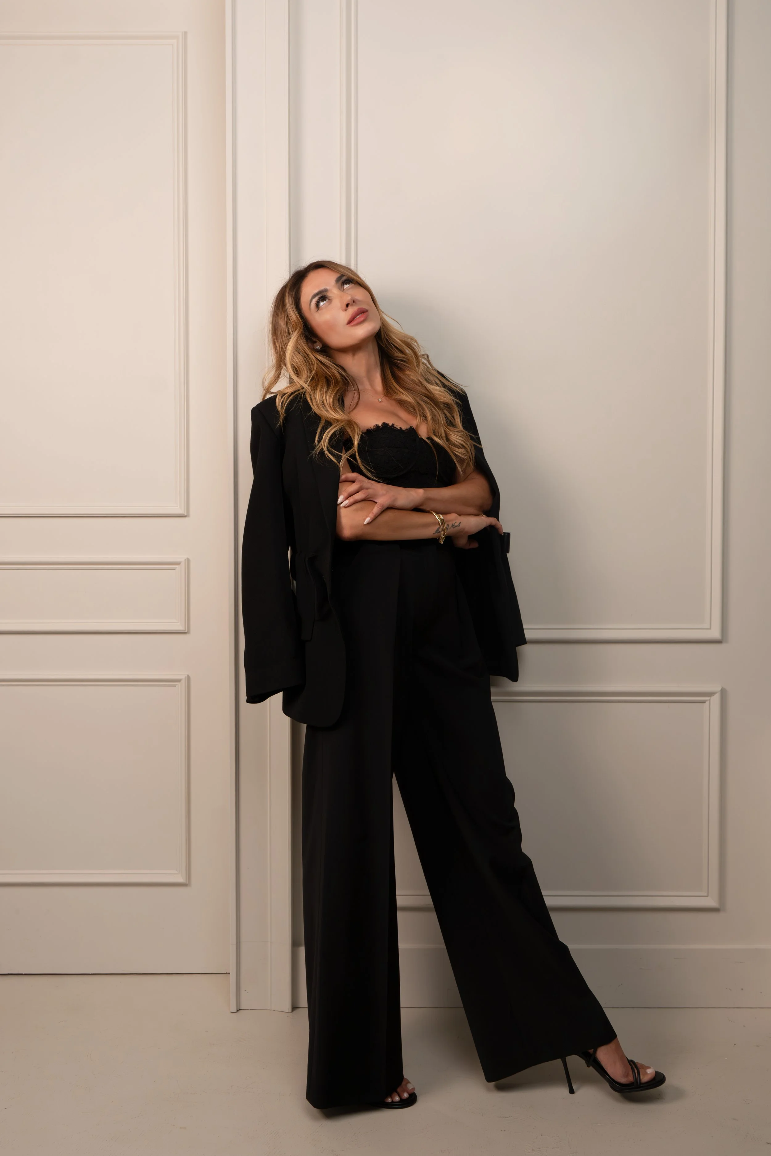 A woman in black pants, black top, and black high heels stands with her arms crossed in front of a white door and wall, looking upwards.