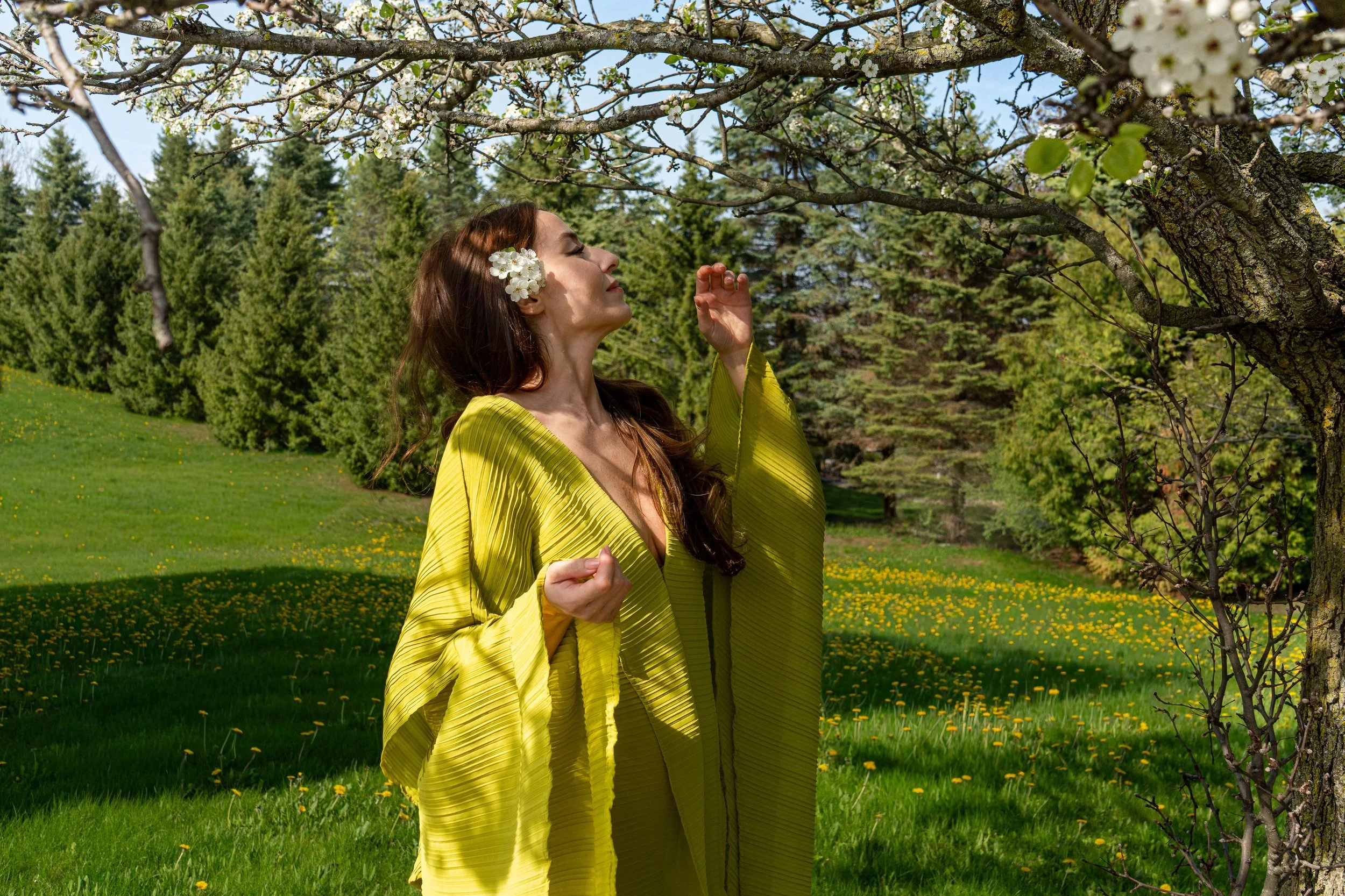 A woman with long brown hair and flowers in her ear wears a yellow pleated dress and stands outdoors under a blossoming tree in a green field with yellow flowers. She has her eyes closed and appears to be enjoying the scent of the flowers.