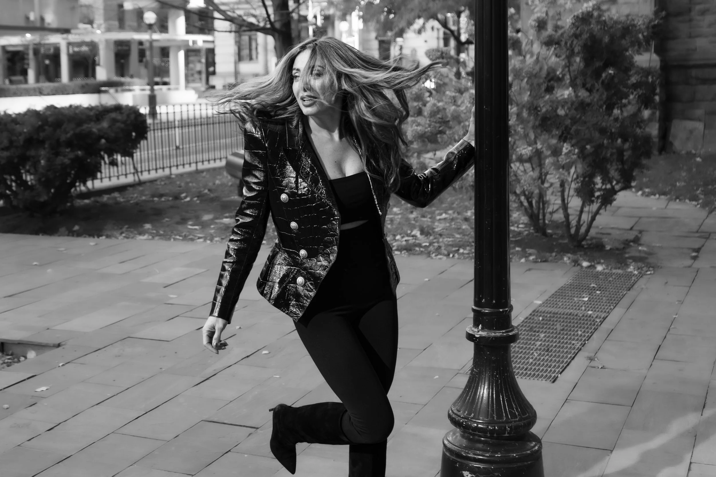 Black and white street fashion image captured mid-motion, wind moving through hair and structured jacket, cinematic urban atmosphere.