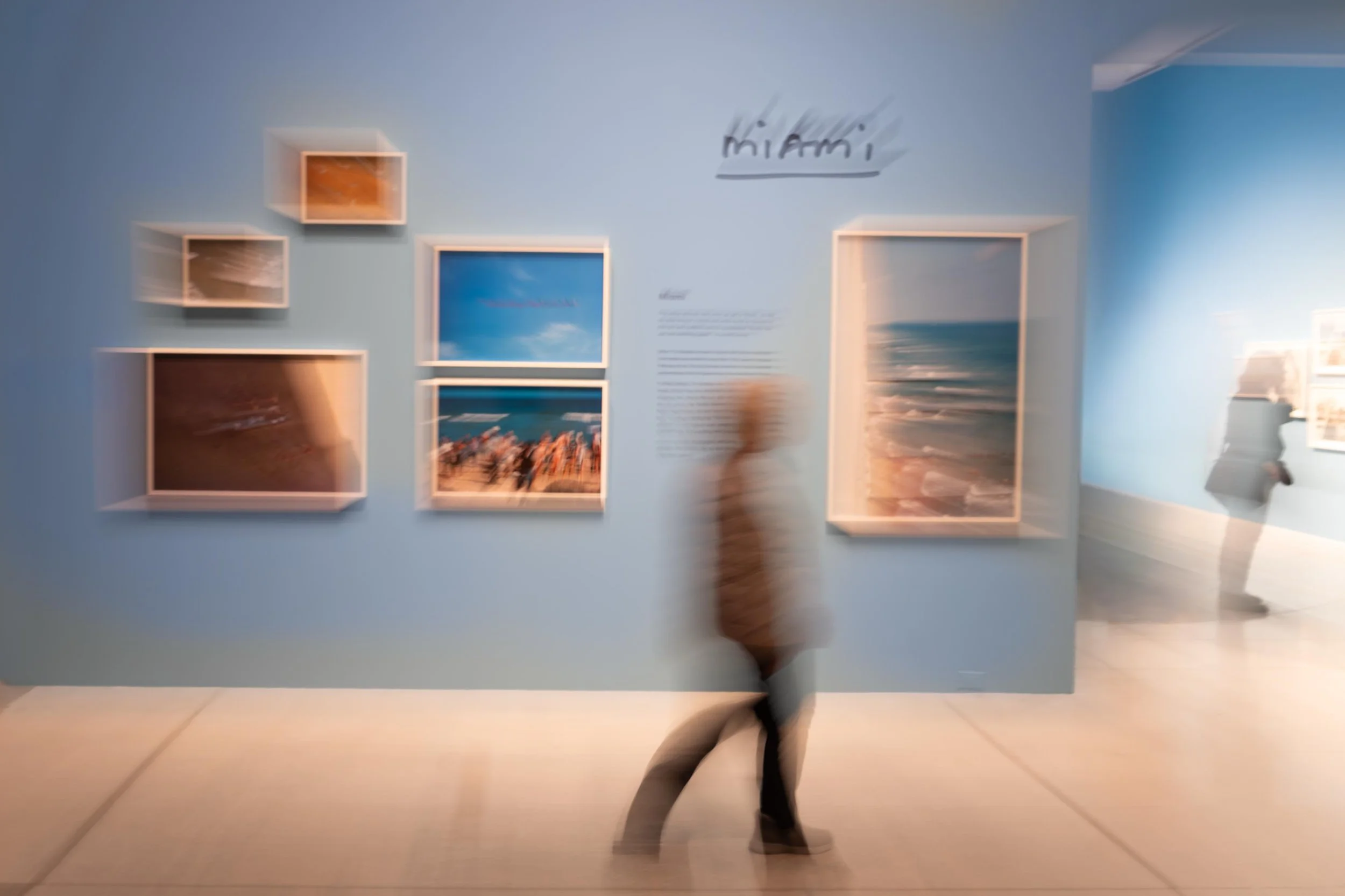 Blurred image of an art gallery with framed pictures on a blue wall and a person walking past