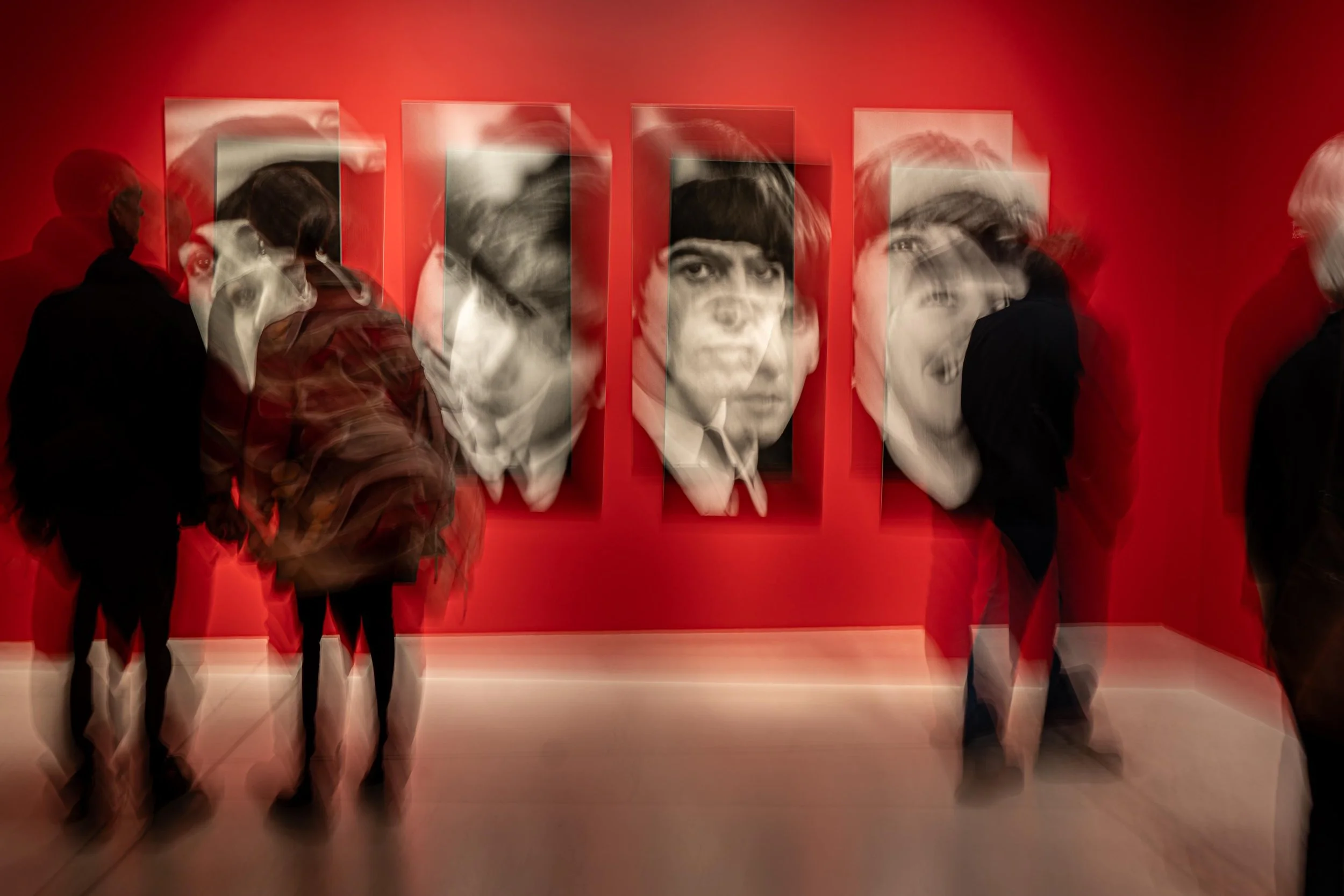 People observing framed portraits of a woman with short hair and a serious expression on a red gallery wall.