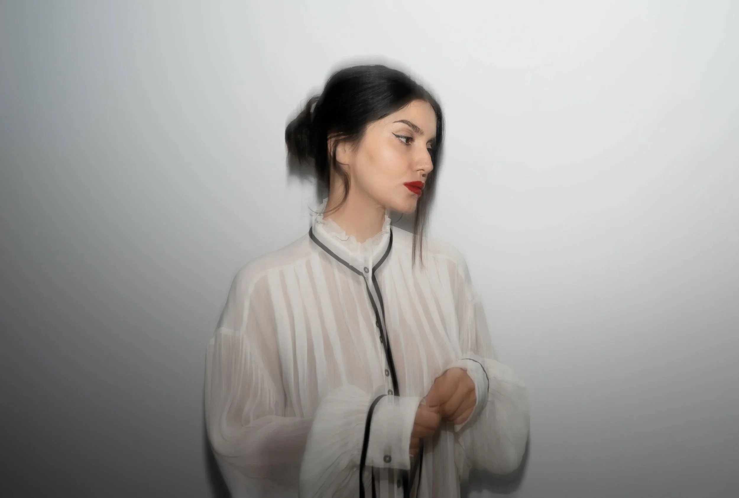A woman with black hair, red lipstick, and winged eyeliner is standing against a plain white wall, wearing a light-colored, vertically striped blouse with black piping along the edges, and is looking downward with her hands clasped.
