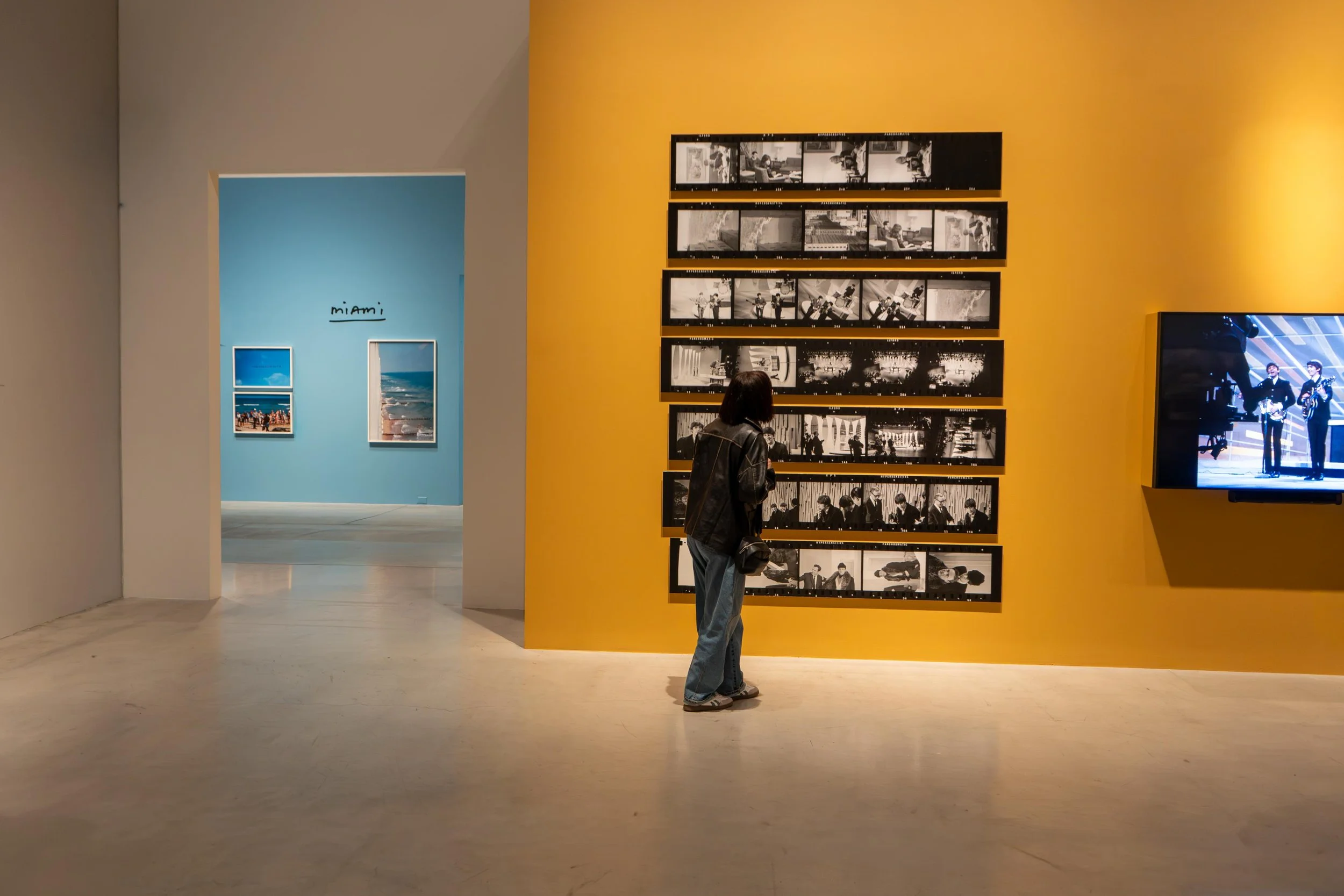 A woman standing and observing black-and-white photographs on a yellow wall in an art gallery. In the background, there is a blue wall with framed pictures and the word 'miami' written on it.