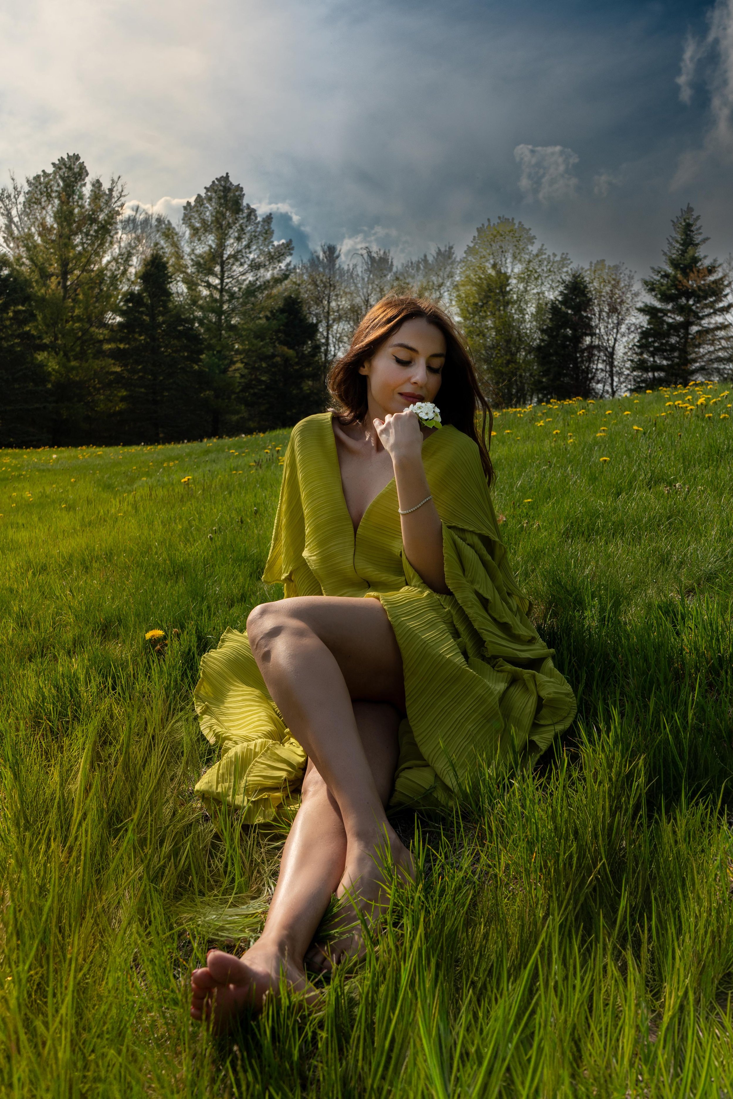 A woman sitting on grass in a field holding a small bouquet of white flowers near her face, with trees and a cloudy sky in the background.