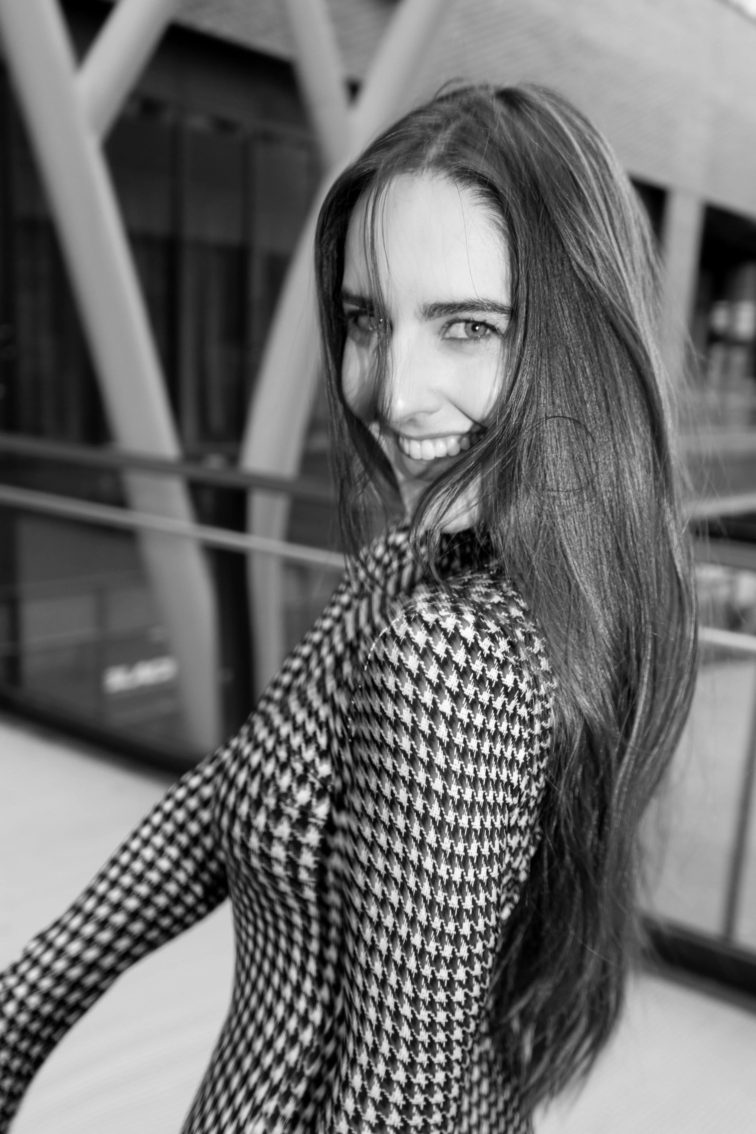 A young woman with long hair and light-colored eyes, smiling while looking over her shoulder. She is wearing a checkered coat, and her hair is partly covering her face. The background appears to be an indoor area with modern architectural elements.