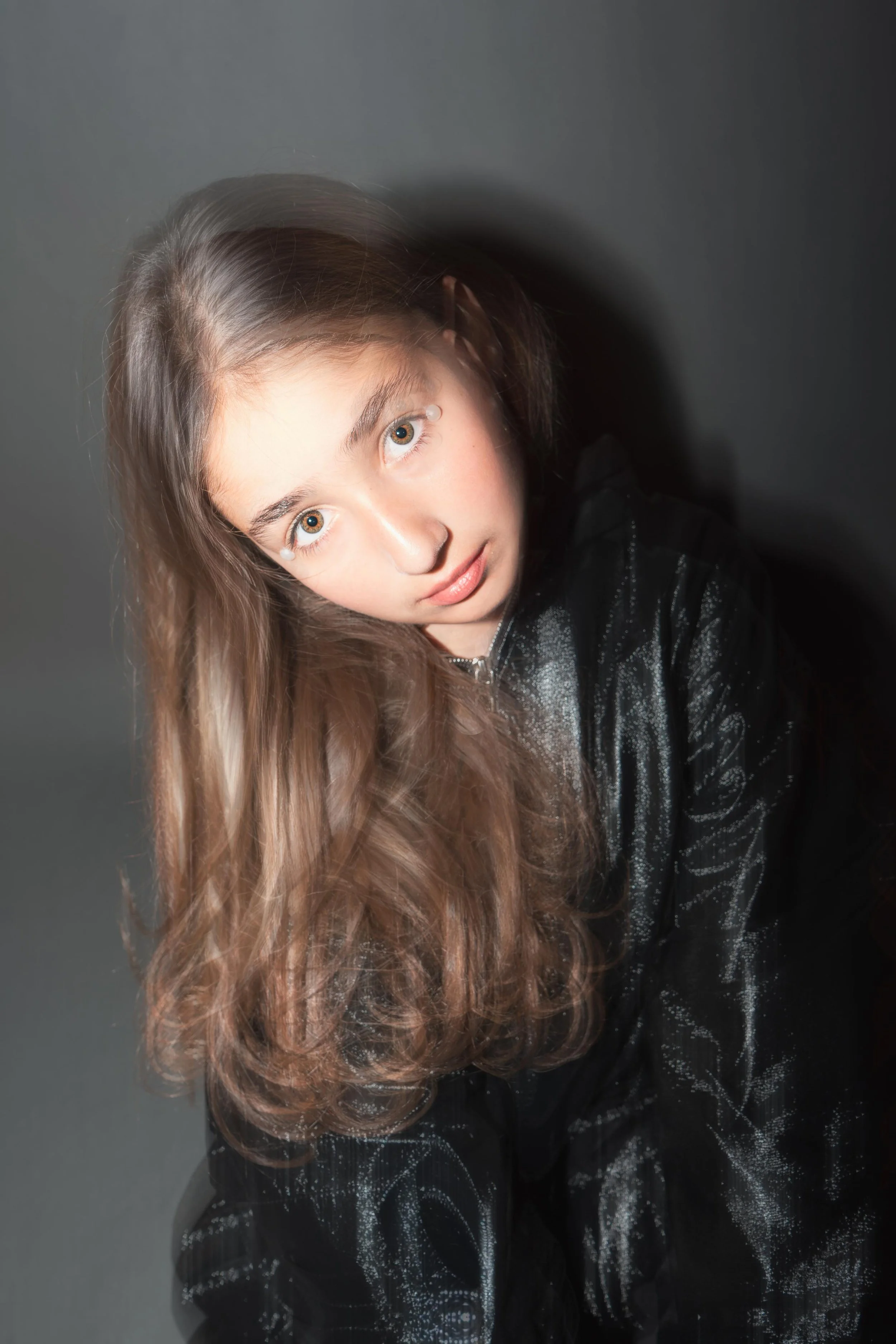 A young girl with long, wavy, brown hair, wearing a black, shiny, textured jacket, looking directly at the camera against a dark, neutral background.
