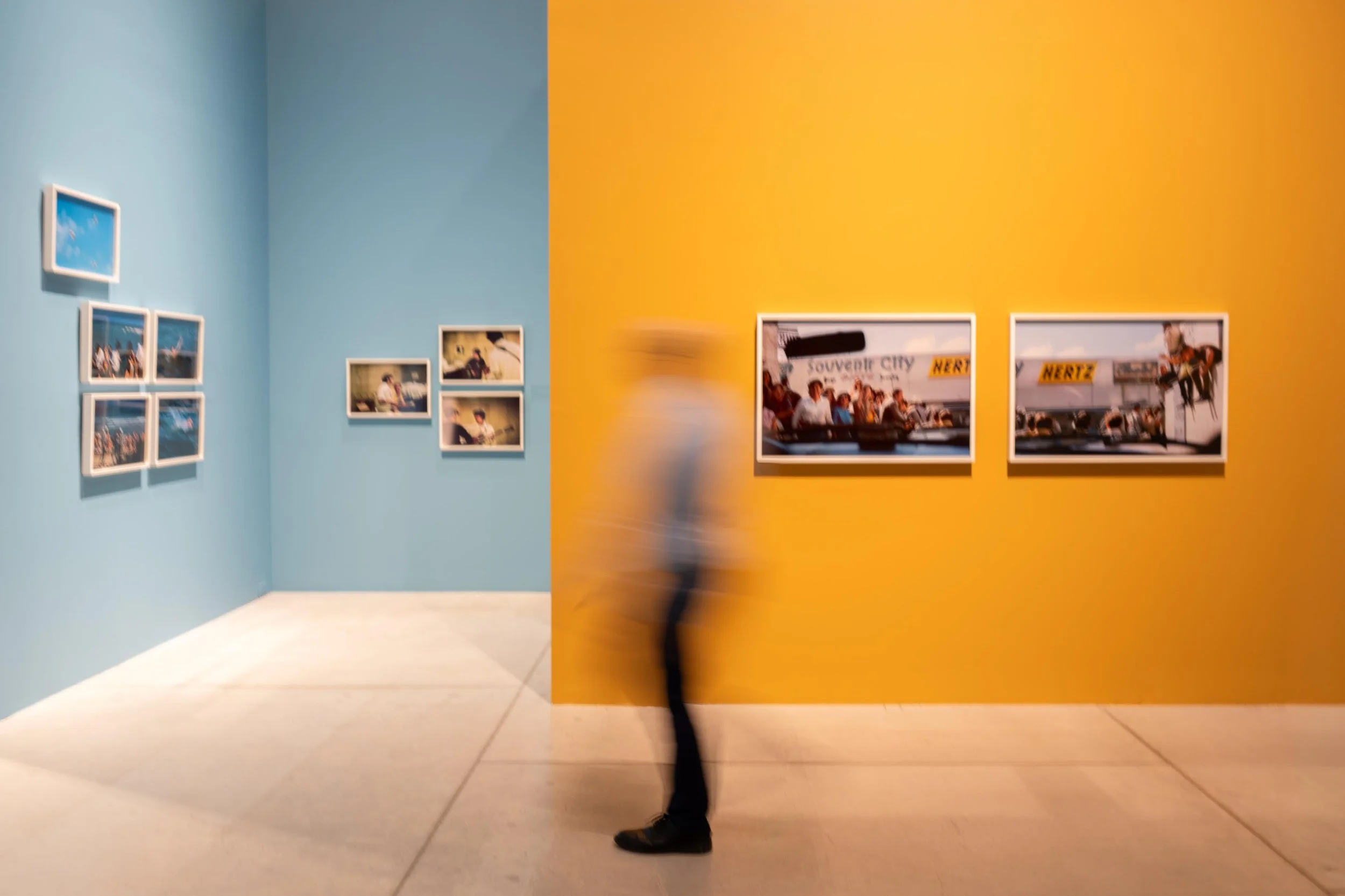 A person walking past an art gallery wall with framed photographs; the wall is blue on the left side and yellow on the right side, and the person is blurred in motion.