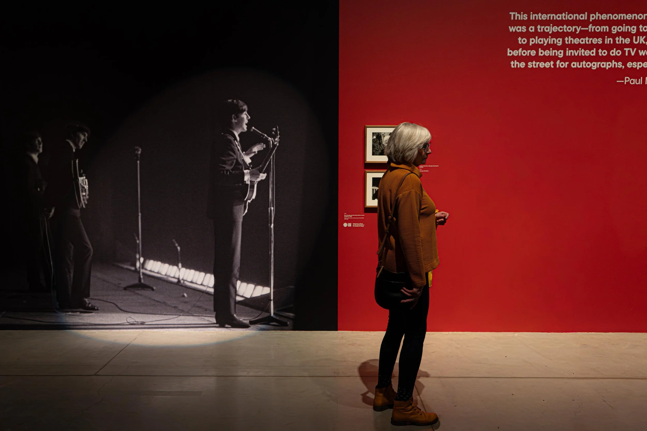 A woman with gray hair wearing an orange striped top and yellow boots is standing in an art gallery, observing a large black-and-white photograph of a man speaking into a microphone on stage. The gallery wall behind her is painted red with text about an international phenomenon in white font, and a smaller framed black-and-white photograph is visible.