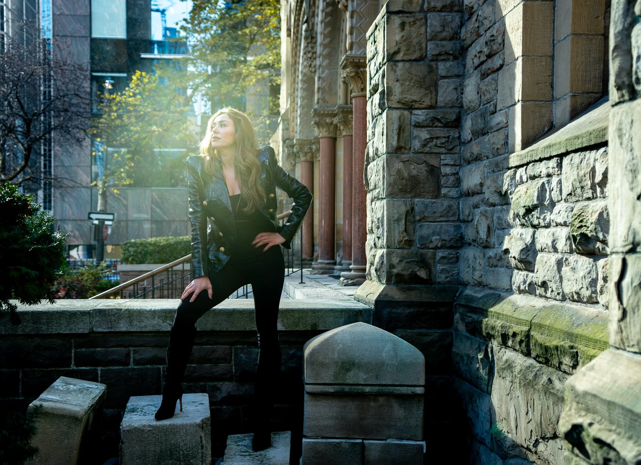 Editorial fashion portrait against historic stone architecture, dramatic light and shadow emphasizing posture, texture, and modern styling.
