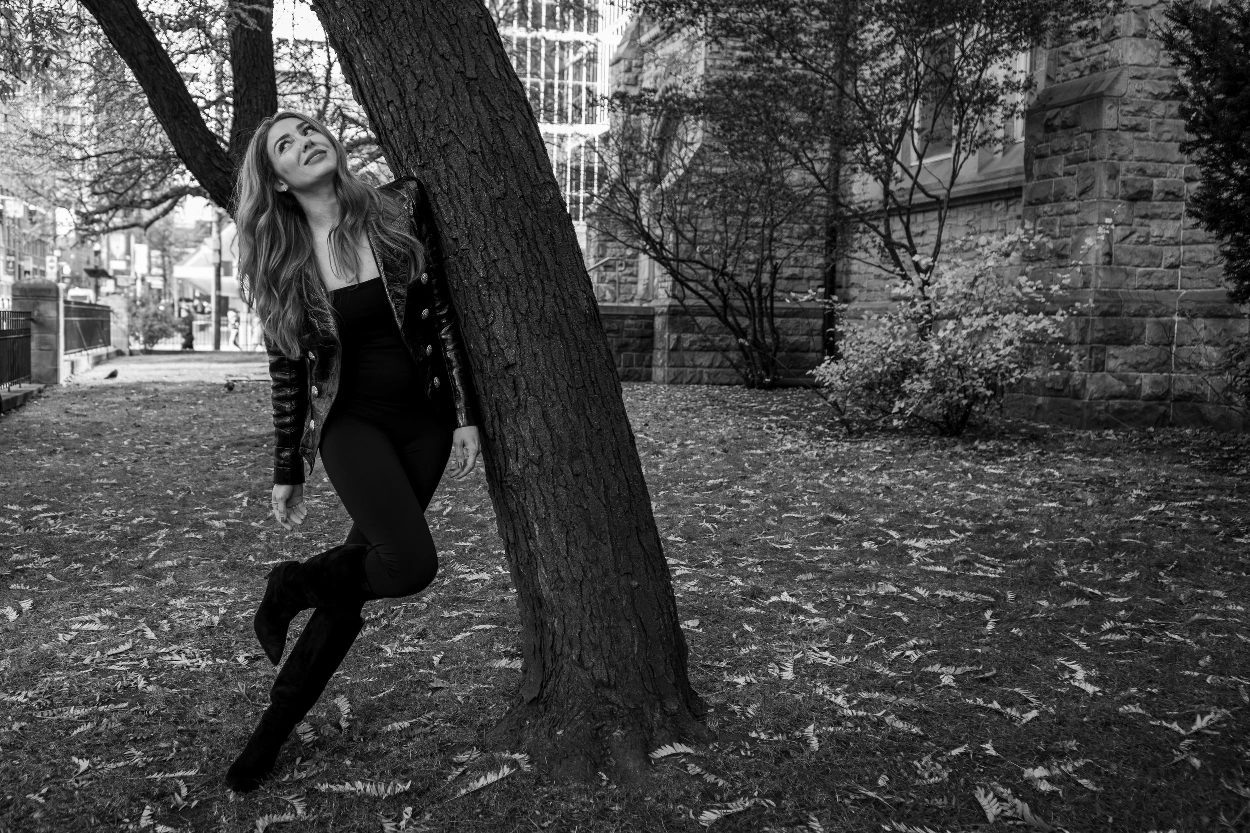 Monochrome outdoor portrait of a woman leaning against a tree in a city park, blending fashion styling with natural textures and movement.