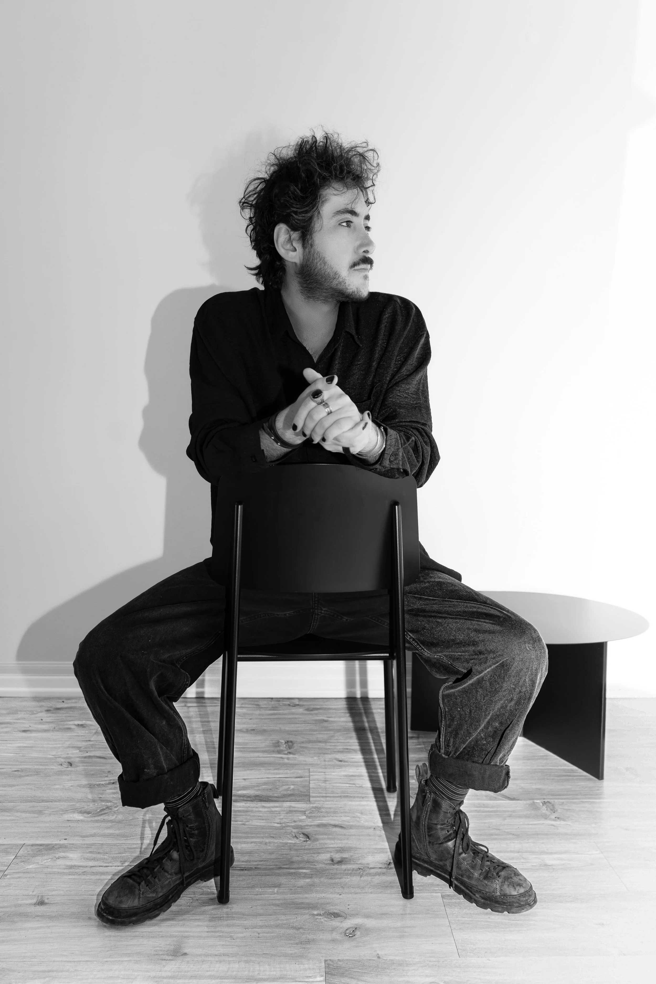A young man with curly hair and a beard wearing a dark shirt, black jeans, and combat boots sitting on a chair with legs spread apart, leaning forward with his hands clasped, looking to the right against a plain white wall, in a black and white photo.