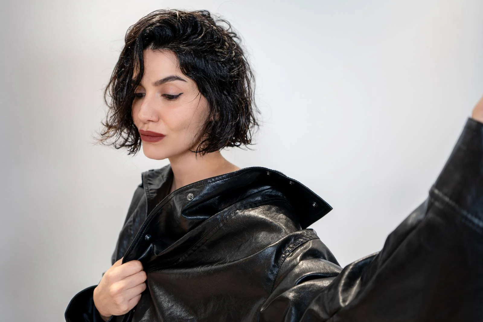 A woman with short, curly black hair and light skin, wearing a black leather jacket, looking down with closed eyes against a plain, light background.