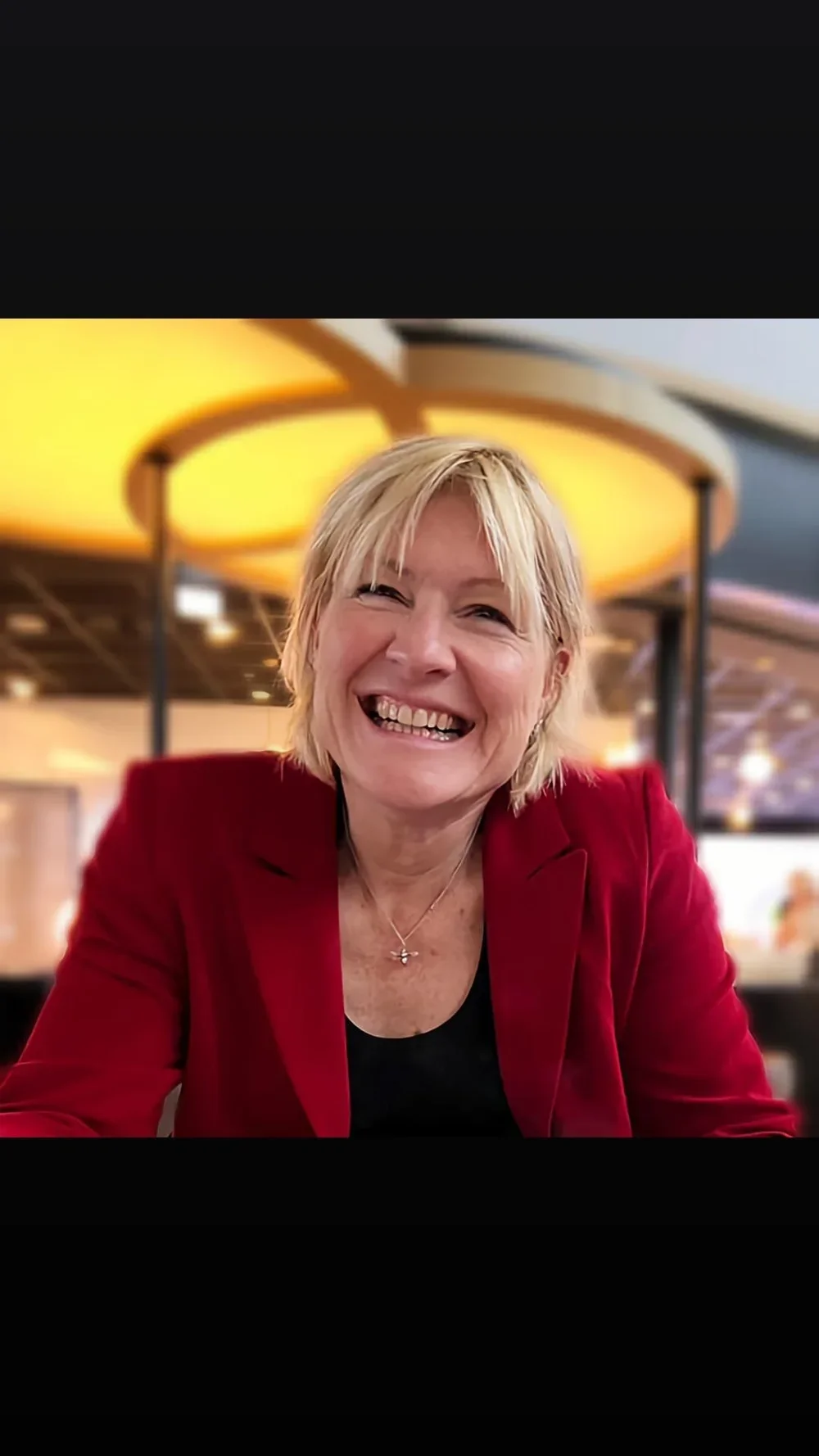 A woman with blonde hair smiling, wearing a red jacket and a black top, in a well-lit indoor setting.