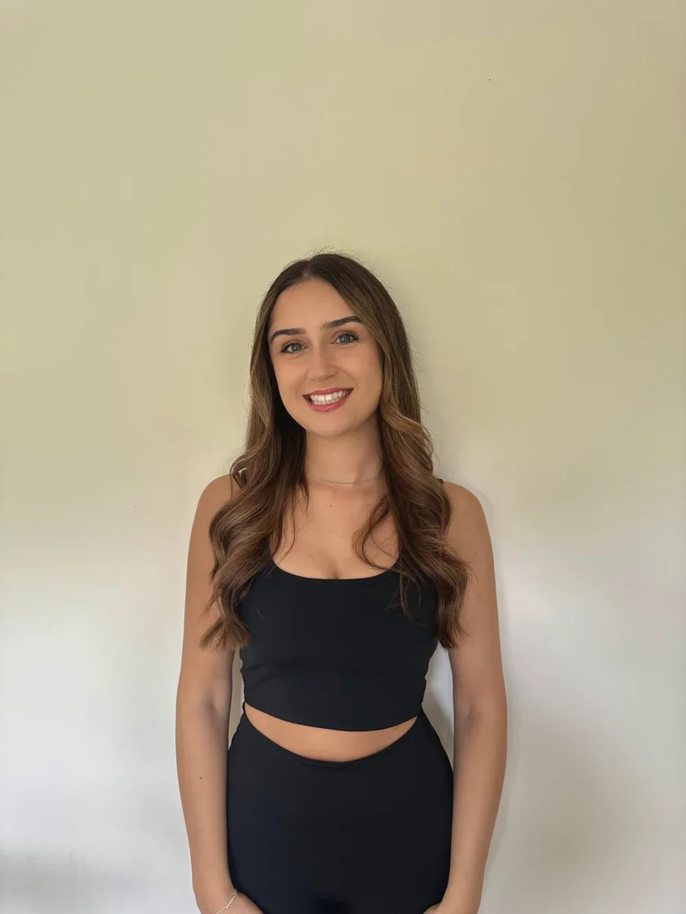 Smiling young woman with long wavy brown hair and fair skin, wearing a black crop top and matching skirt, standing against a plain beige wall.