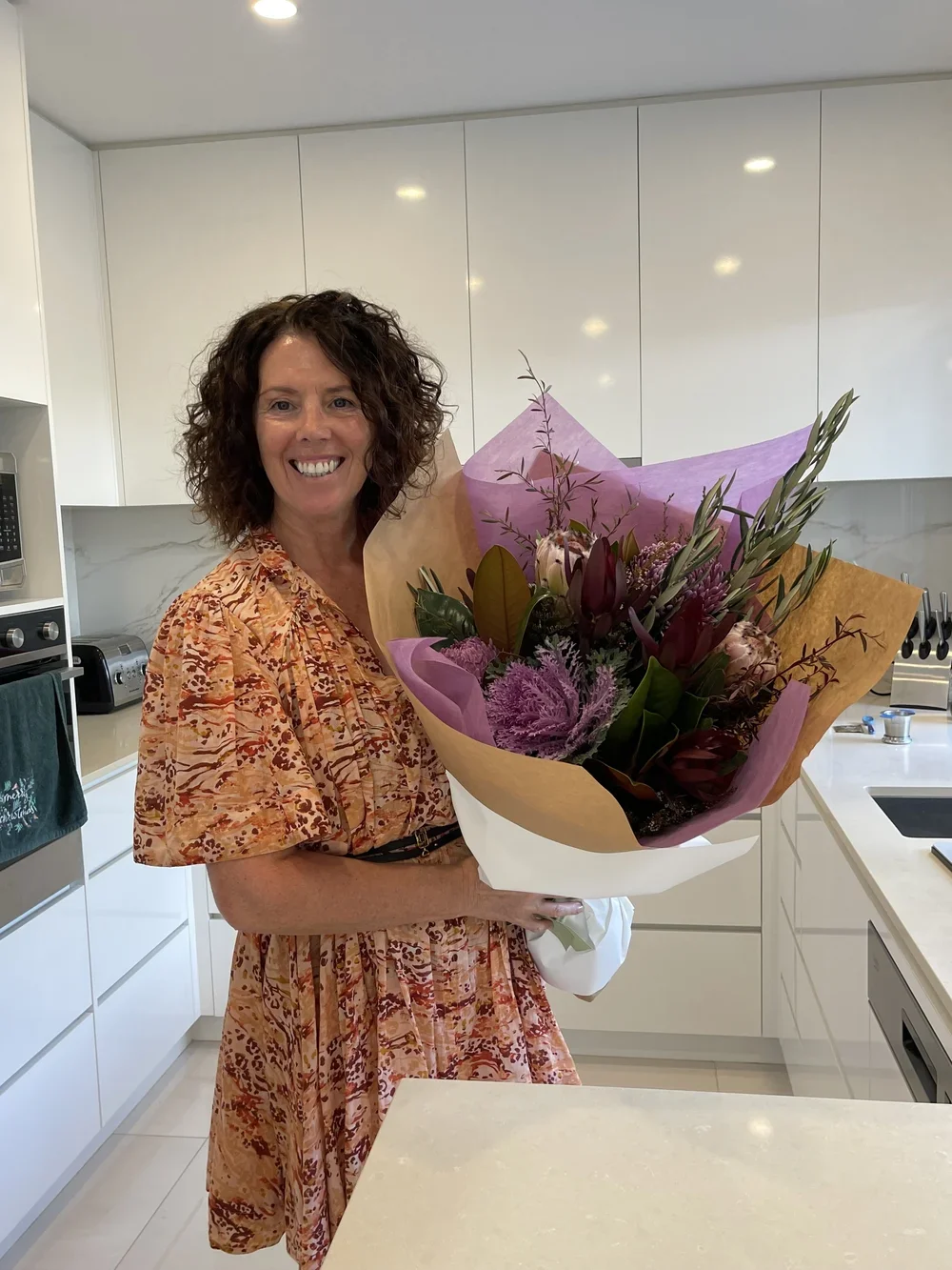A woman with curly brown hair smiling and holding a large bouquet of pink and purple flowers in a modern white kitchen.