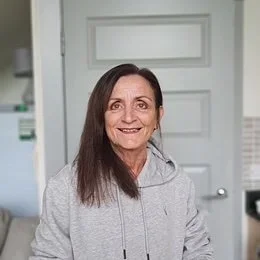 A woman with long brown hair wearing a gray hoodie stands indoors, smiling at the camera.