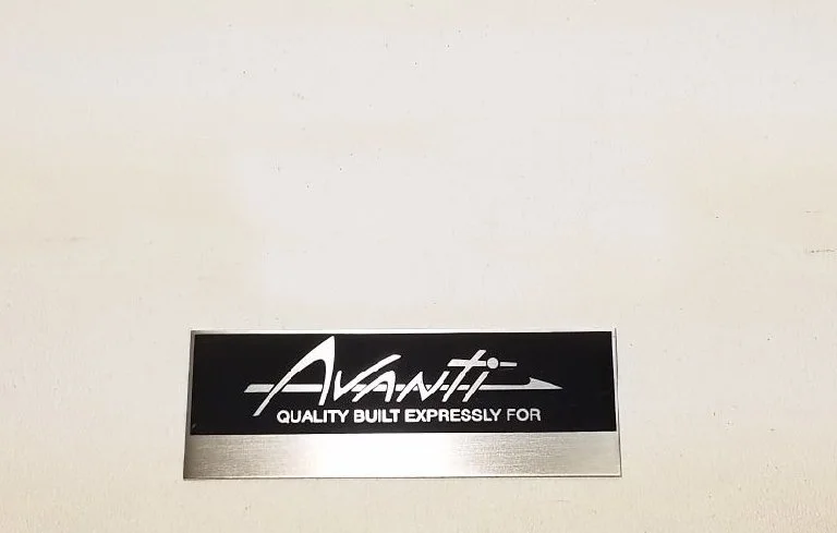Genuine factory original Avanti dash plaque without stripes, original peel-and-stick backing, 3.5 inches wide.