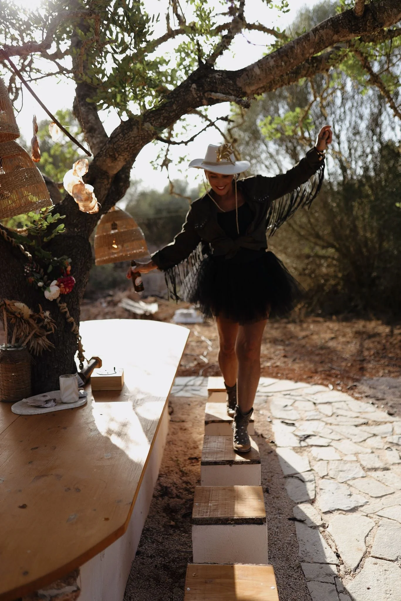Nadine Miree  in einem weißen Hut und dunkler Kleidung balanciert auf einem Holzplanke, die auf einem Wandersockel steht, im Freien unter einem Baum, im Hintergrund sind Sonnenlicht und Bäume sichtbar.