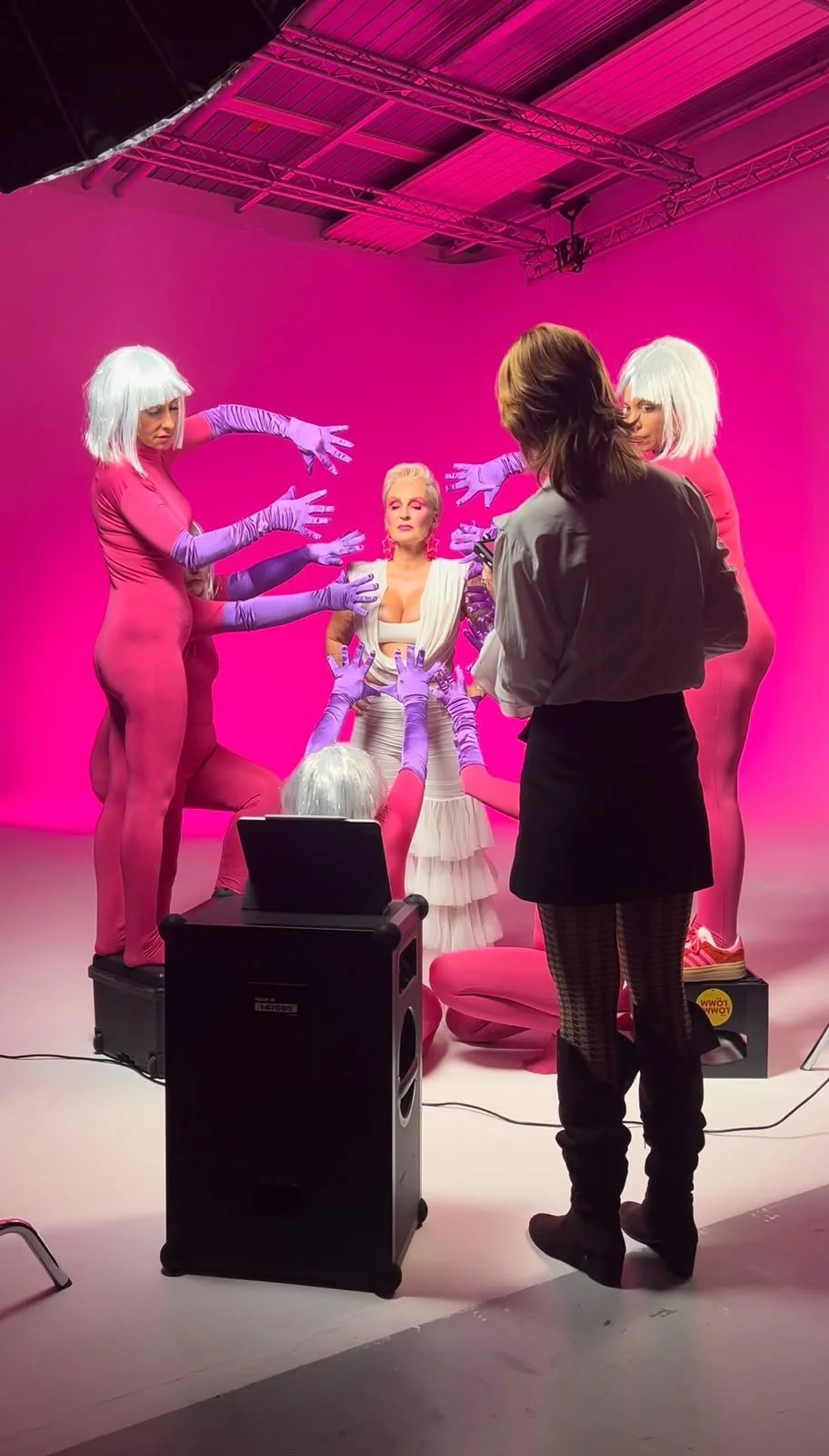 Fotoshooting in einem pink beleuchteten Studio mit fünf Frauen, drei in pinken Ganzkörperanzügen, eine in weißen Kleid, eine fotografiert. Die Frauen in Anzügen tragen weiße Perücken, die andere hat braune Haare. Es stehen Scheinwerfer und Technik im Studio.