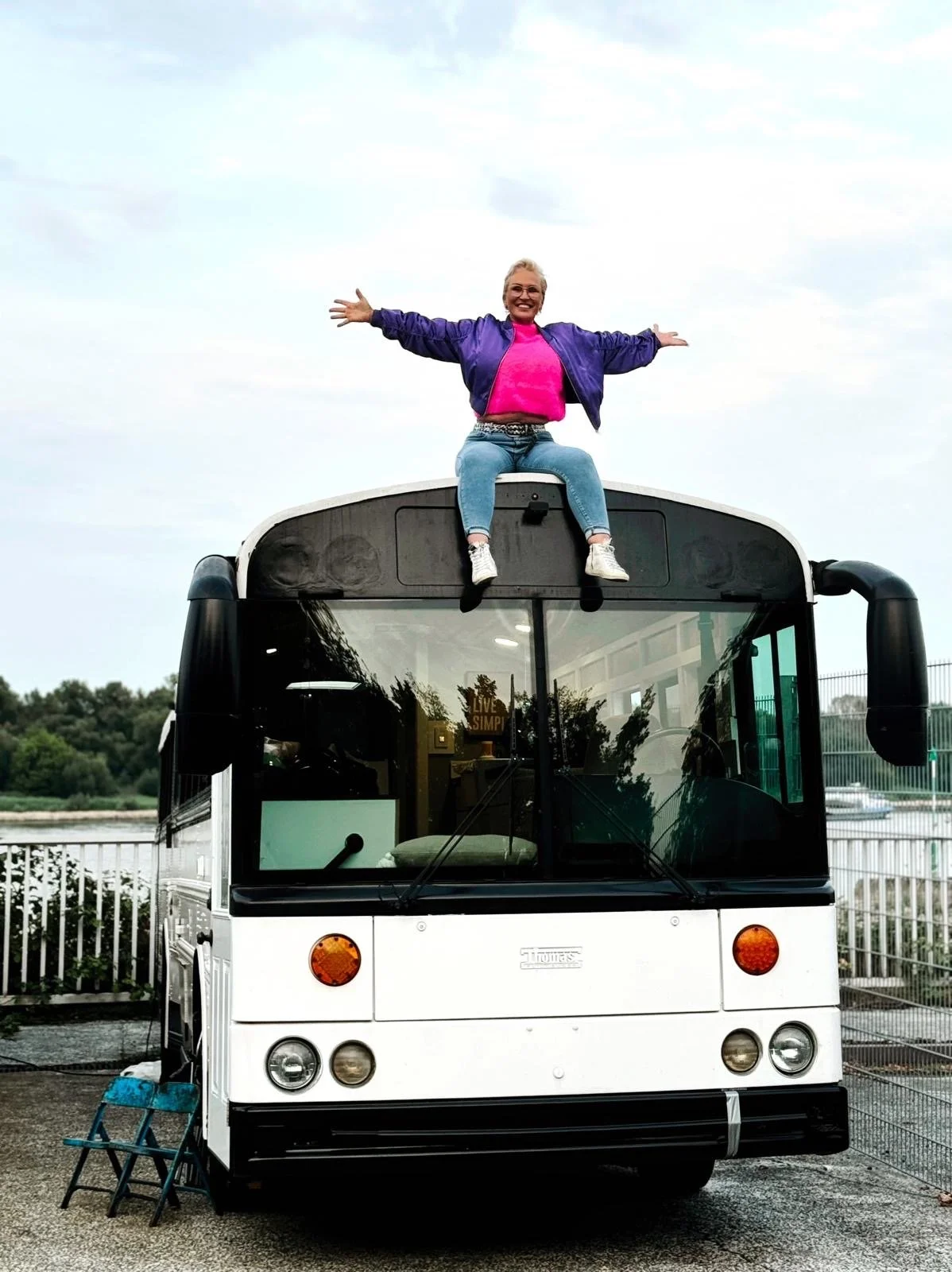 Nadine Miree sitzt auf dem Dach eines weißen Buses (Schulbus Lukie) mit offenen Armen. Im Hintergrund sind Bäume und ein Himmel mit Wolken.