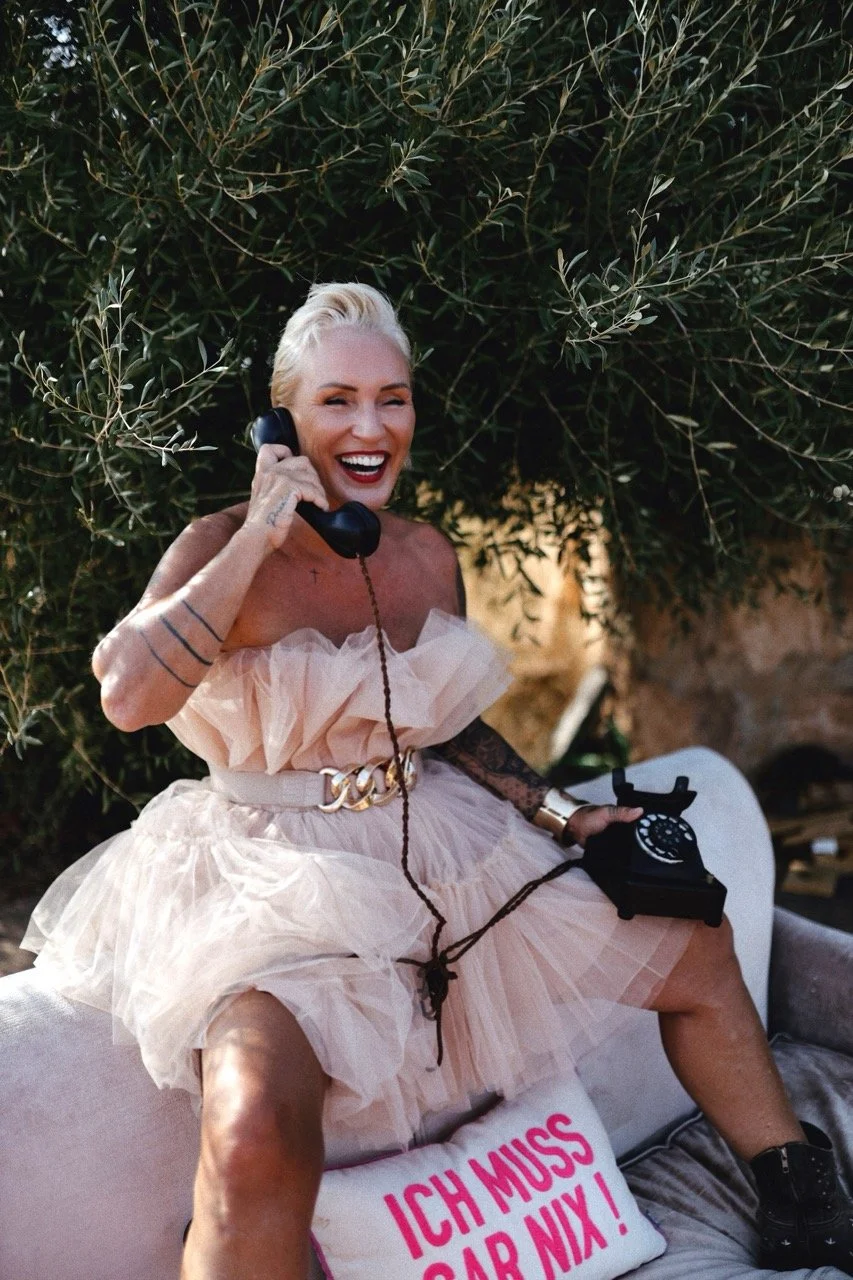 Nadine Miree  sitzt auf einem Sofa, hält ein altes Telefon und trägt ein elegantes, pastellfarbenes Kleid, umgeben von Pflanzen im Freien.