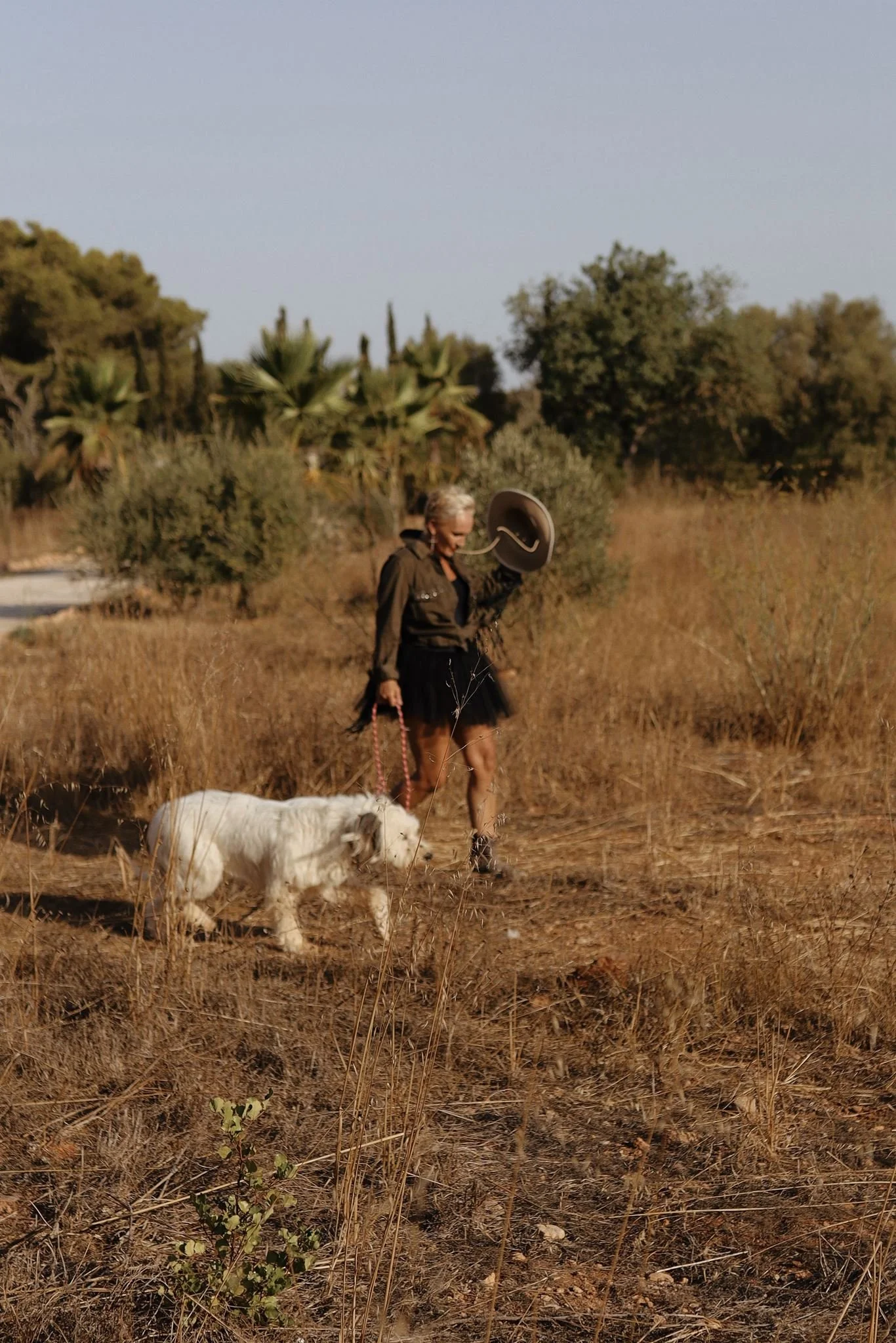 Eine Frau mit grauen Haaren und dunkler Kleidung geht mit einem weißen Hund in einem trockenen Grasland, während sie einen Hut in der Hand hält.