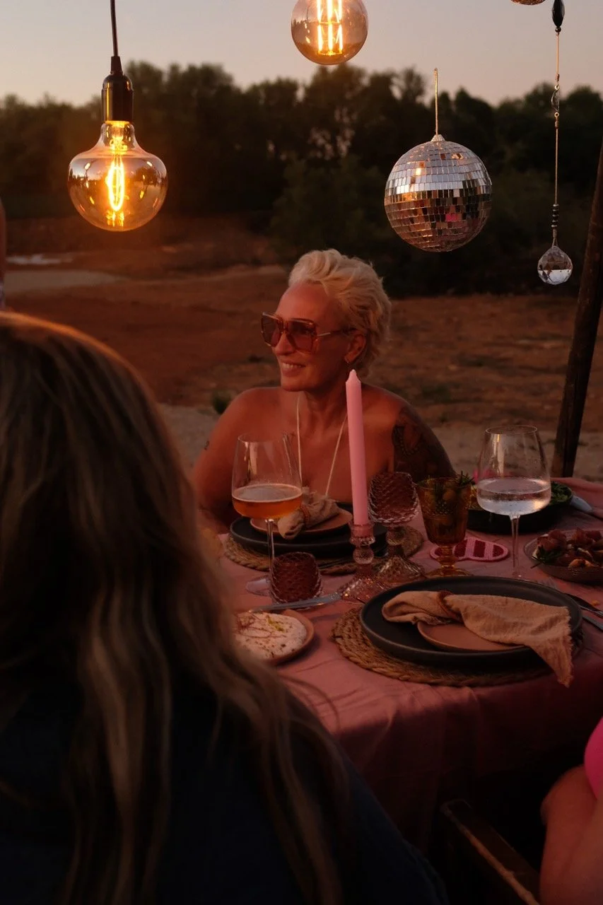 Eine Frau mit kurzen blonden Haaren und Sonnenbrille sitzt an einem Esstisch bei Sonnenuntergang im Freien, umgeben von dekorativen Lichterketten und einem Disco-Kugel, die eine festliche Atmosphäre schaffen.