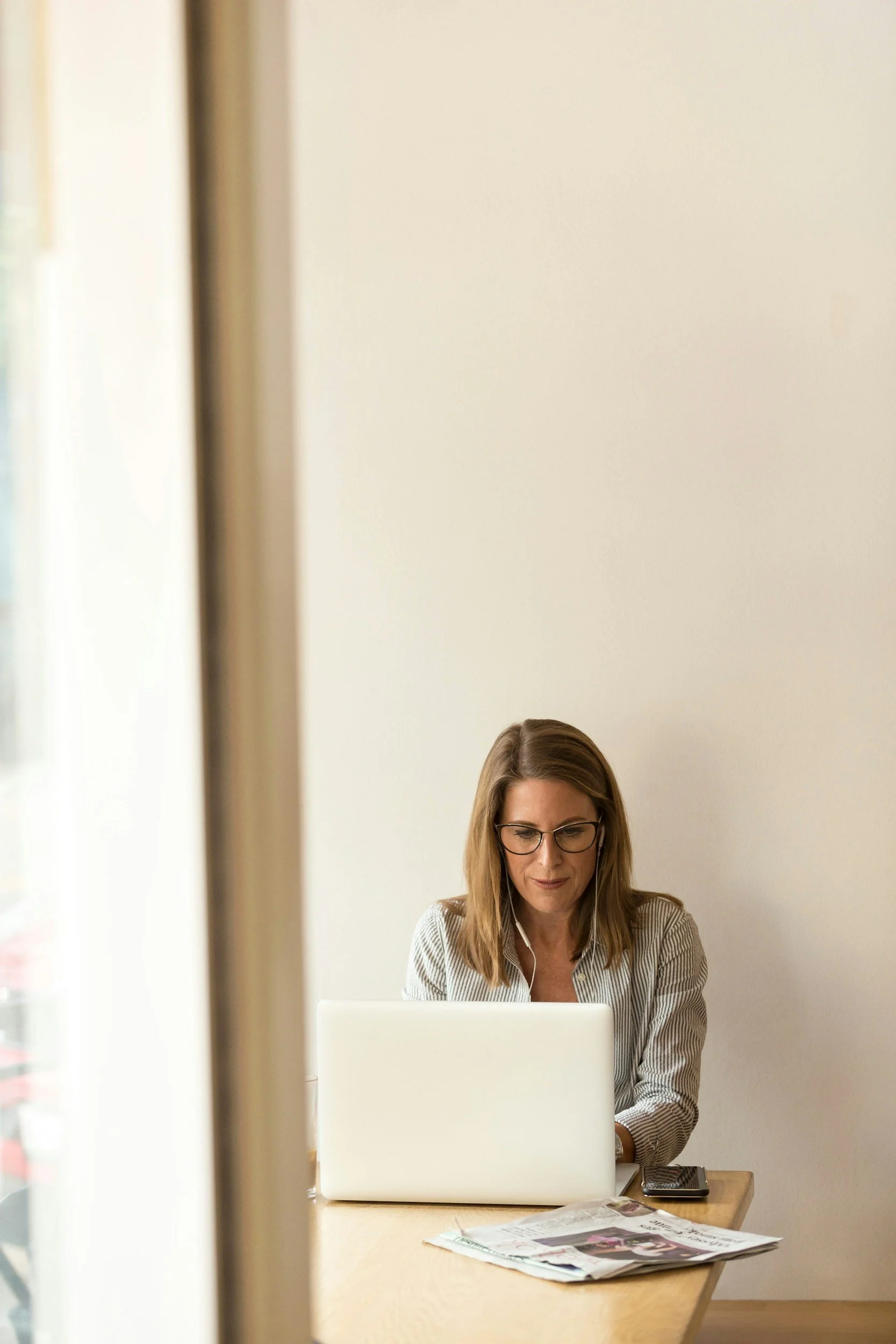 A woman with shoulder-length brown hair and glasses sitting at a wooden desk using a laptop, with a newspaper and a phone on the desk, in a room with a plain white wall and a window on the left side.