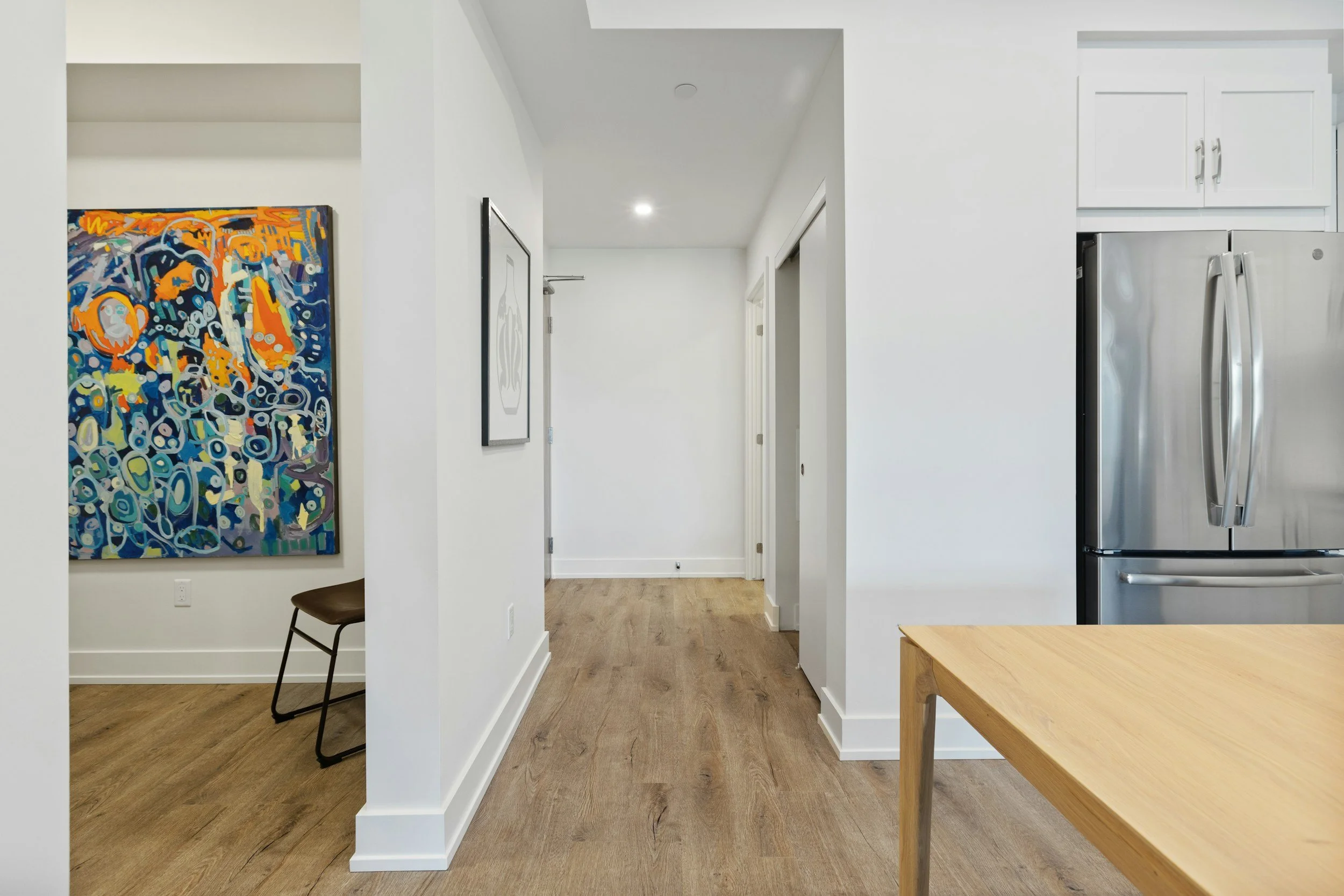Interior view of a modern kitchen with white walls, wood flooring, a stainless steel refrigerator, a light wood table, and artwork on the walls.