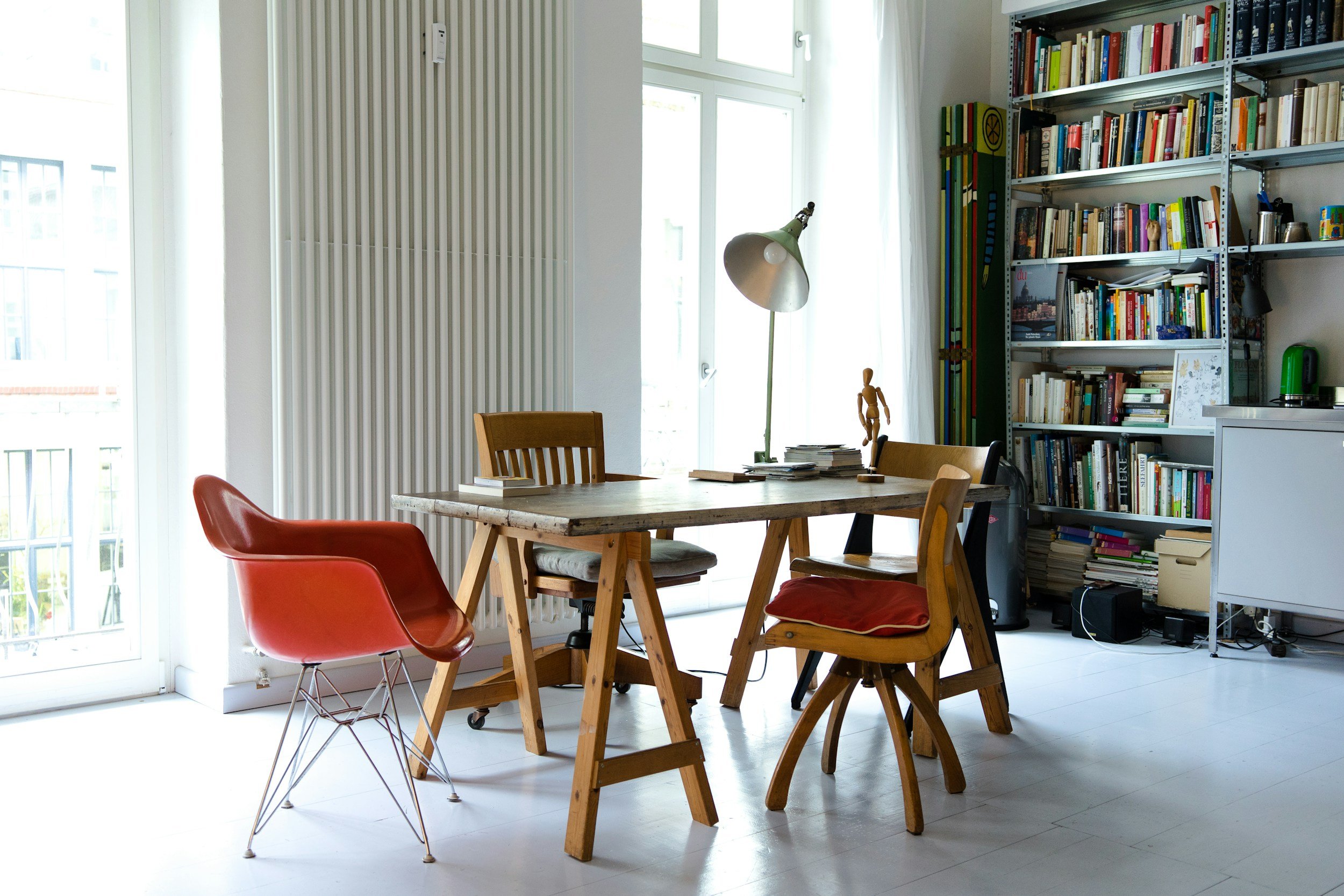 Bright, modern interior with a wooden dining table surrounded by various chairs, a bookshelf filled with books, a large window letting in natural light, and a white cabinet on the right.