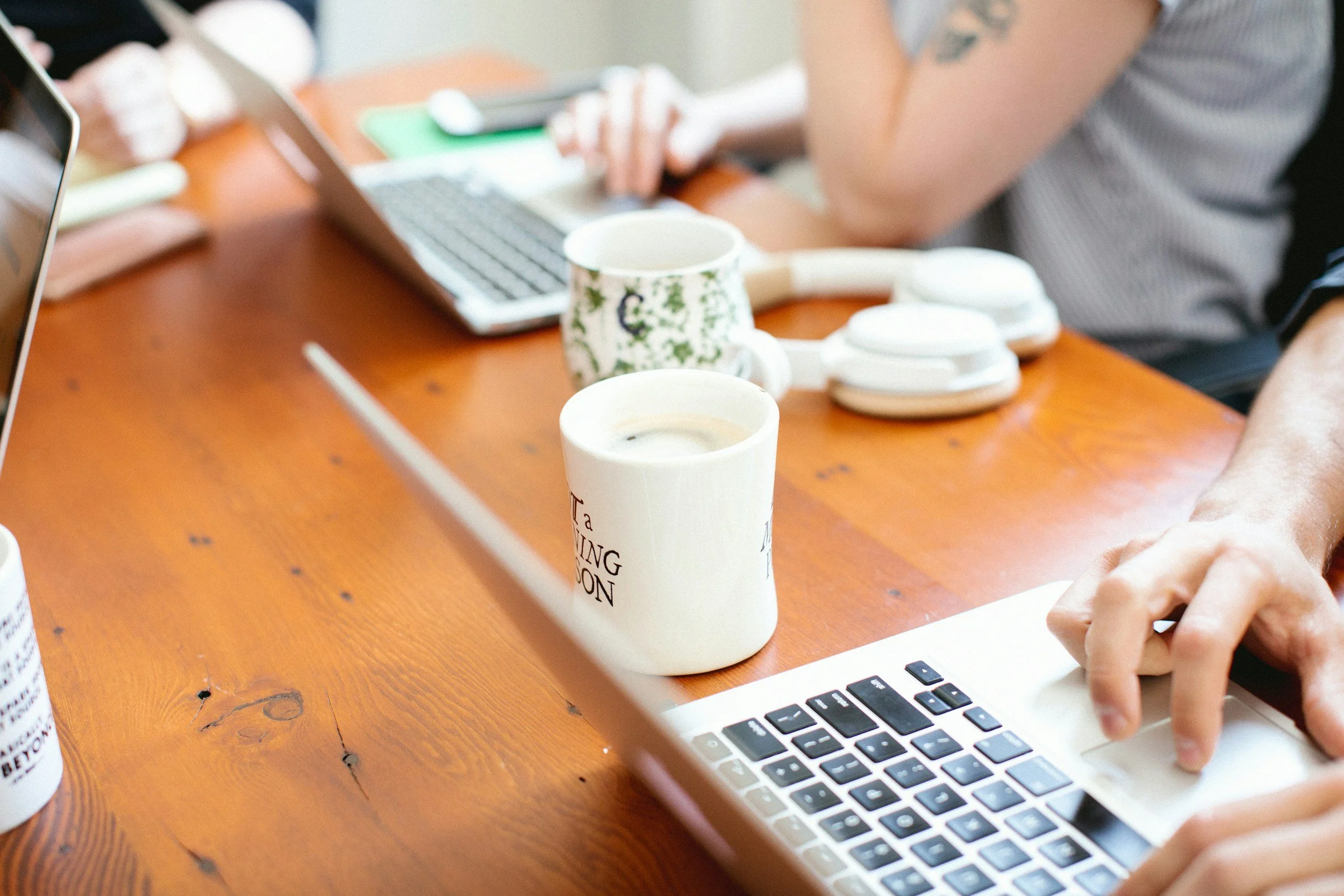 Close-up of a person's hand using a laptop on a wooden table with coffee mugs and headphones.