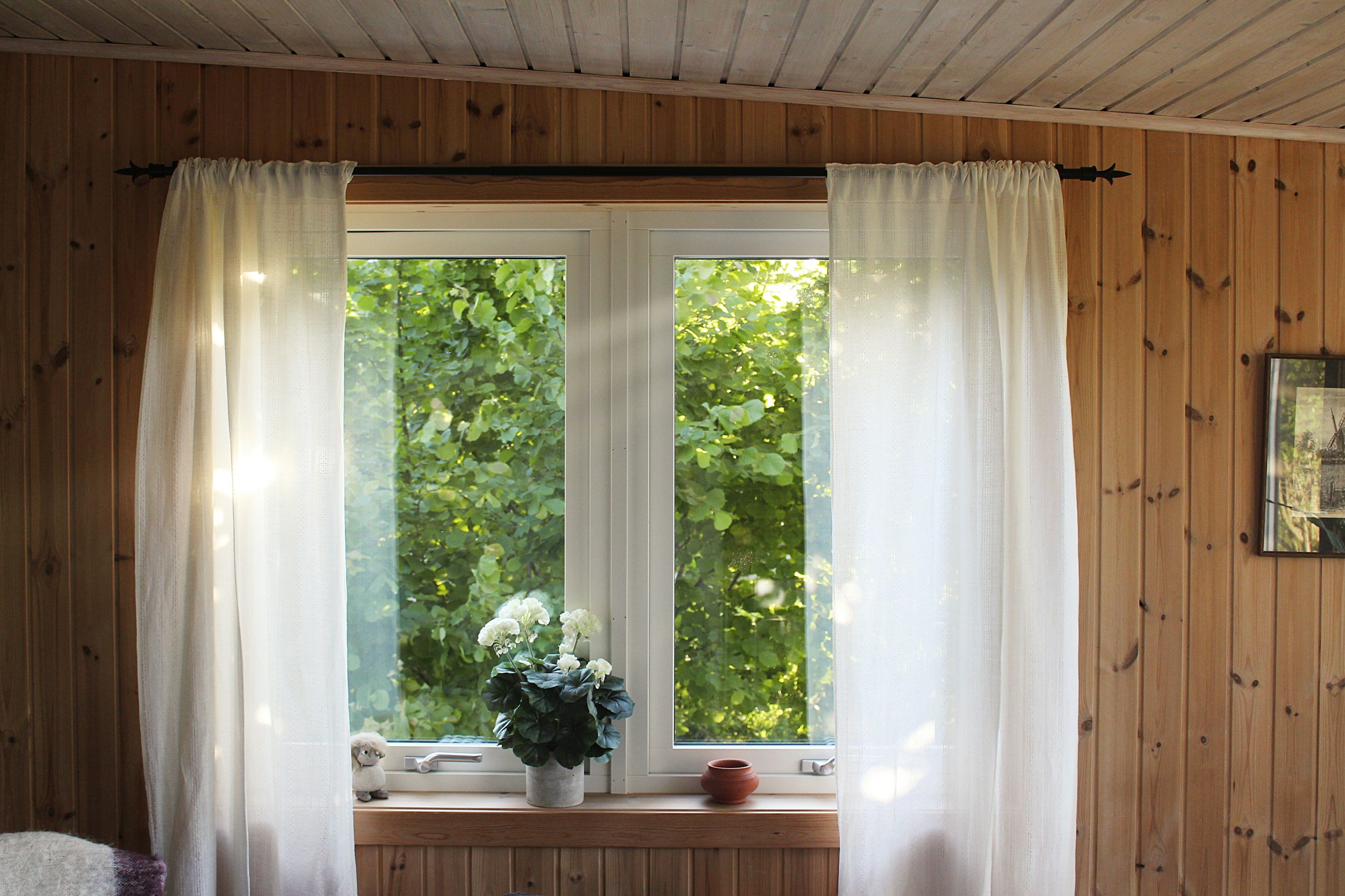 Living room with wooden paneled walls and a large window covered with white curtains, overlooking green trees outside. There are decorative items on the windowsill, including a potted plant with white flowers and a small brown pottery piece.