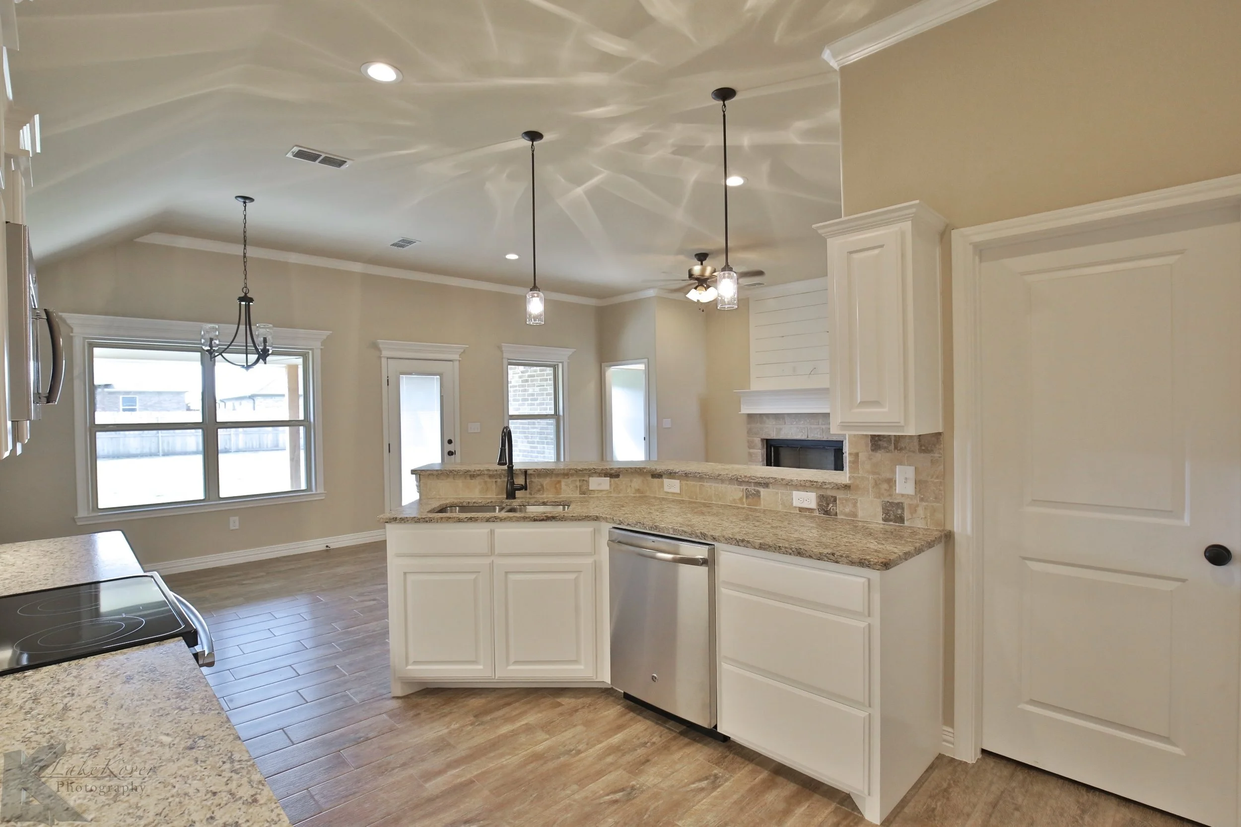 Open-concept kitchen with granite countertops, white cabinets, a stainless steel dishwasher, and a breakfast bar, with a living room featuring a fireplace and multiple windows in the background.
