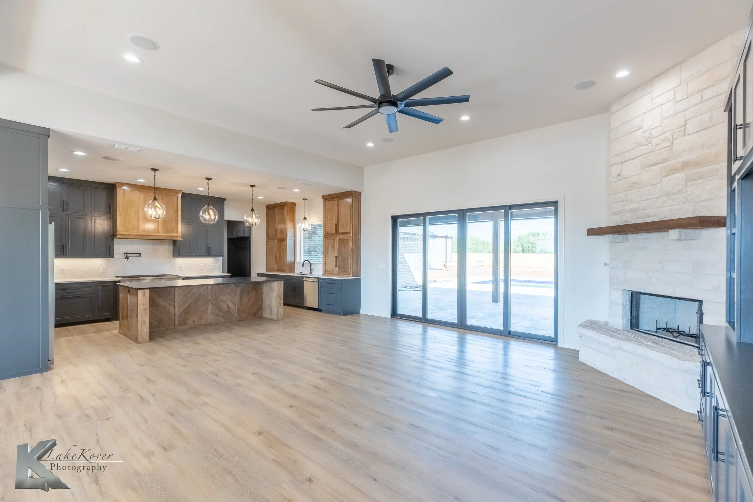 Open concept living area with kitchen, large sliding glass doors, and a fireplace with a wooden mantel. The room features a wood floor, modern ceiling fan, and recessed lighting.