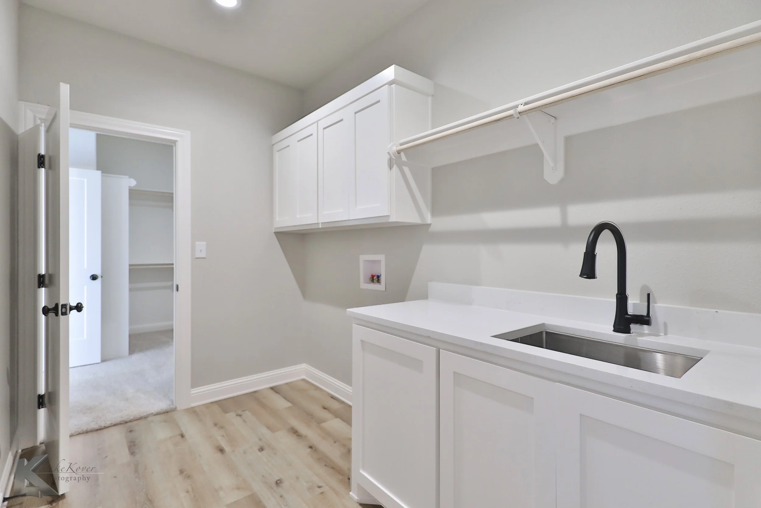 Empty laundry room with white cabinets, a white countertop, a black faucet, a small sink, and a shelf above. An open door reveals a walk-in closet with white shelves.