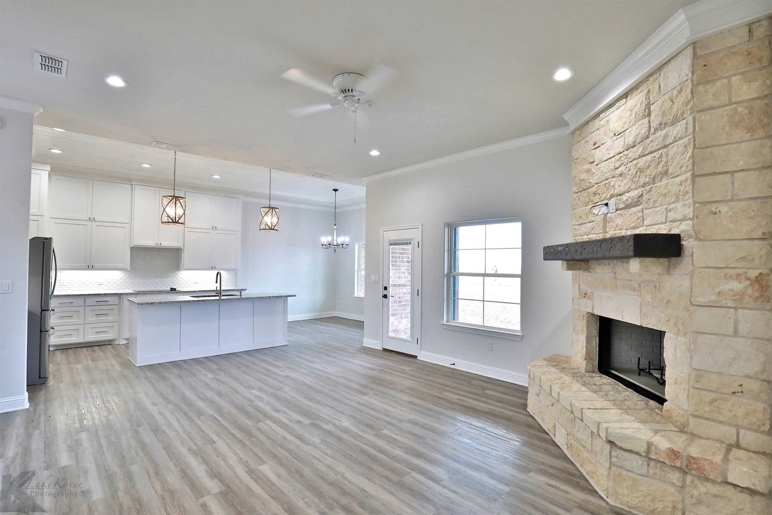 Empty open-concept living room and kitchen area with white cabinetry, a stone fireplace, hardwood floors, a large window, and pendant lighting.