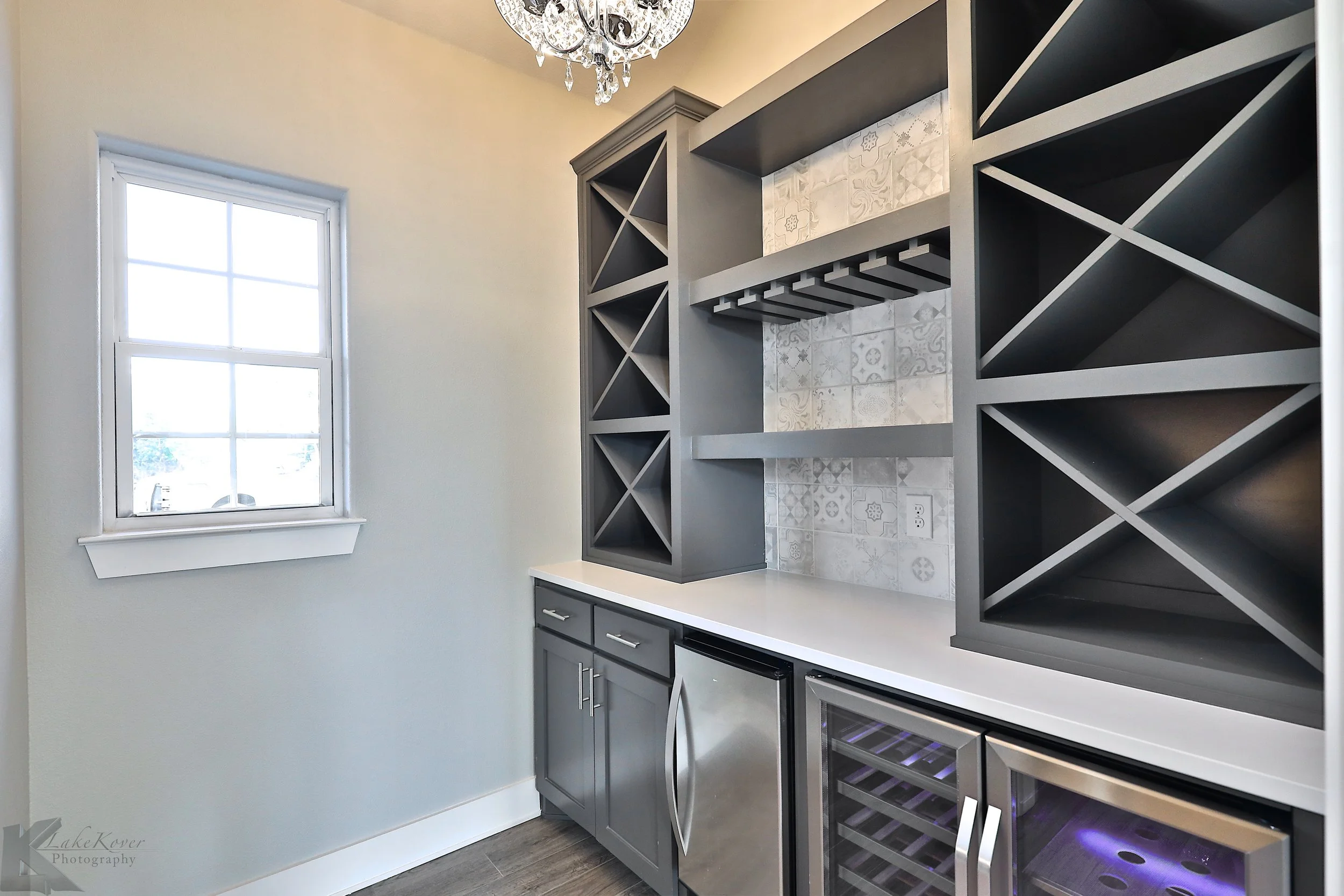 A home bar with gray cabinets, open wine racks above and below, a countertop, and a window to the left. A chandelier hangs from the ceiling.