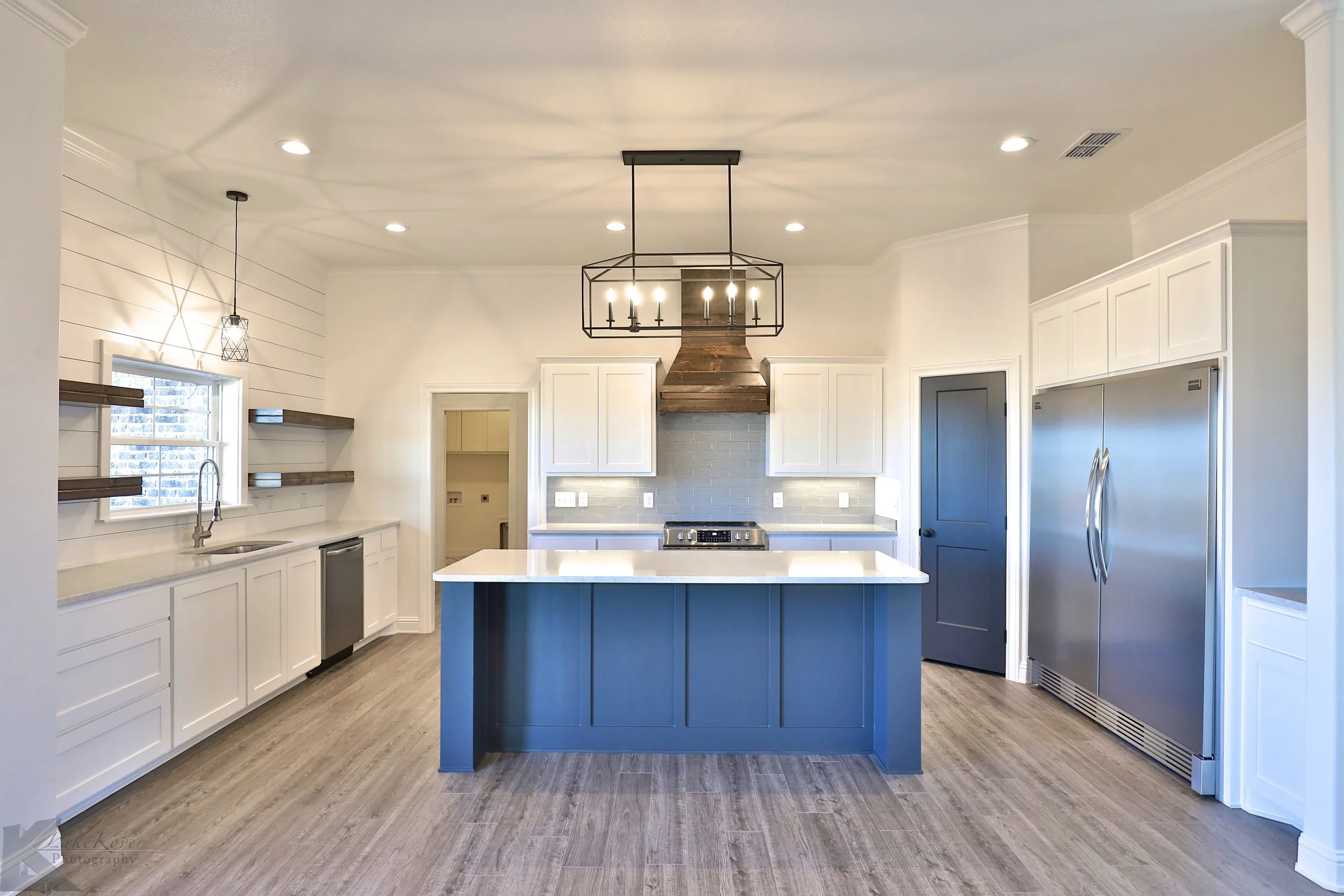 Modern kitchen with white cabinets, a blue island, stainless steel refrigerator, wood shelves, and a gray backsplash, illuminated by ceiling lights and a chandelier.