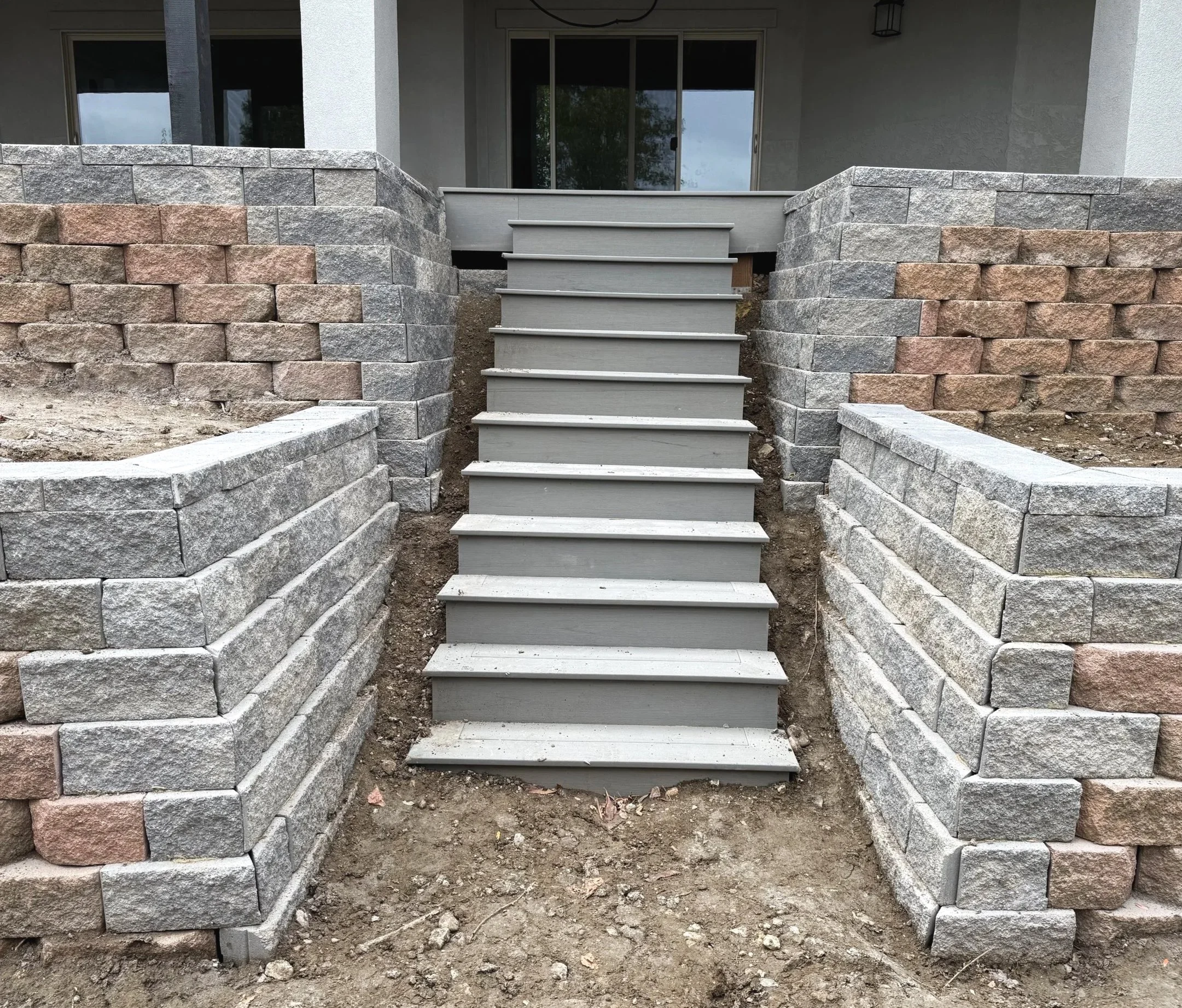 Concrete steps leading up to a house with a sliding glass door and a window, flanked by stone retaining walls on both sides, with exposed dirt and construction materials in the foreground.