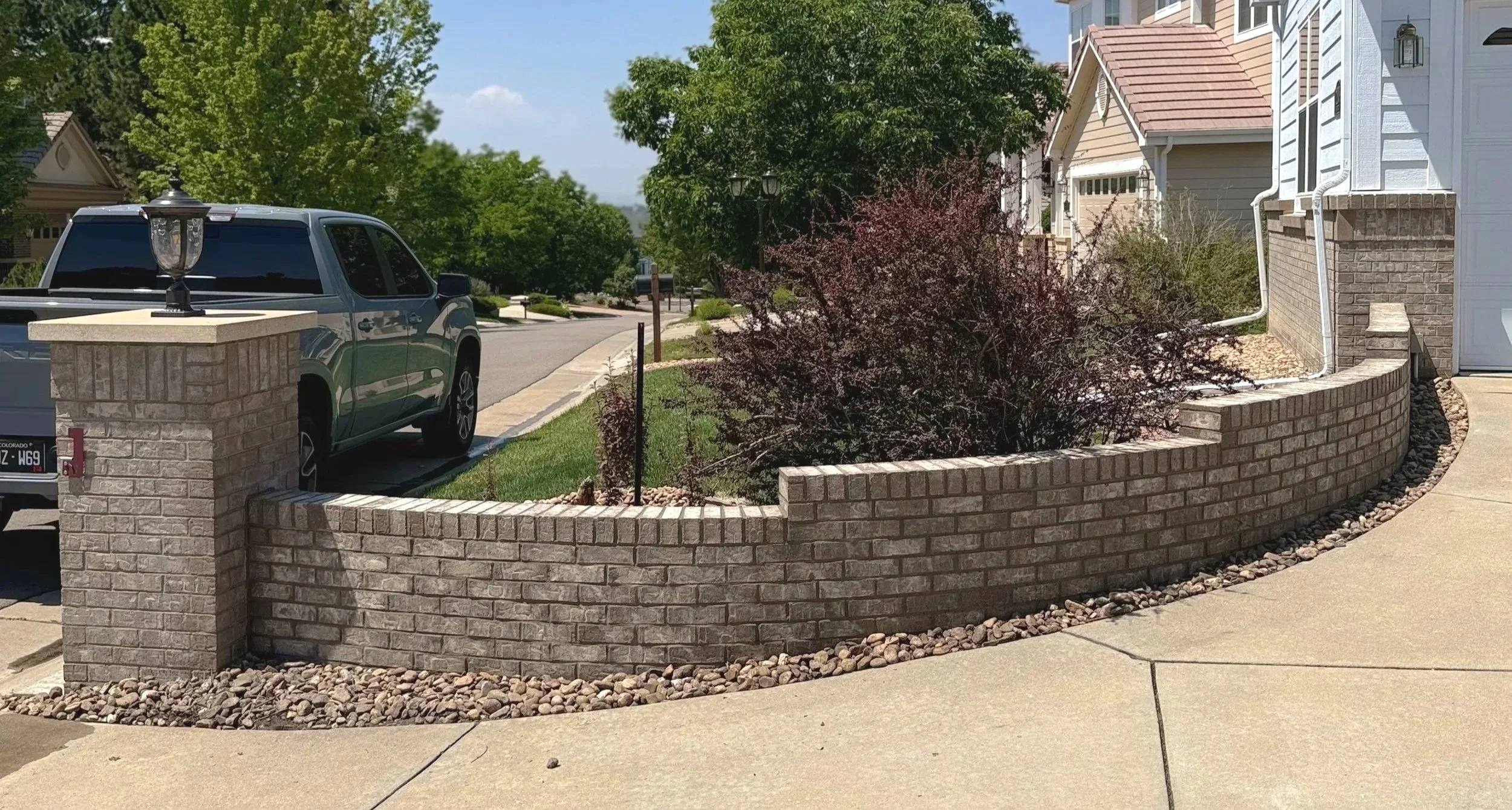A residential street view with a brick wall, a parked truck, a sidewalk, trees, and houses with colorful siding.