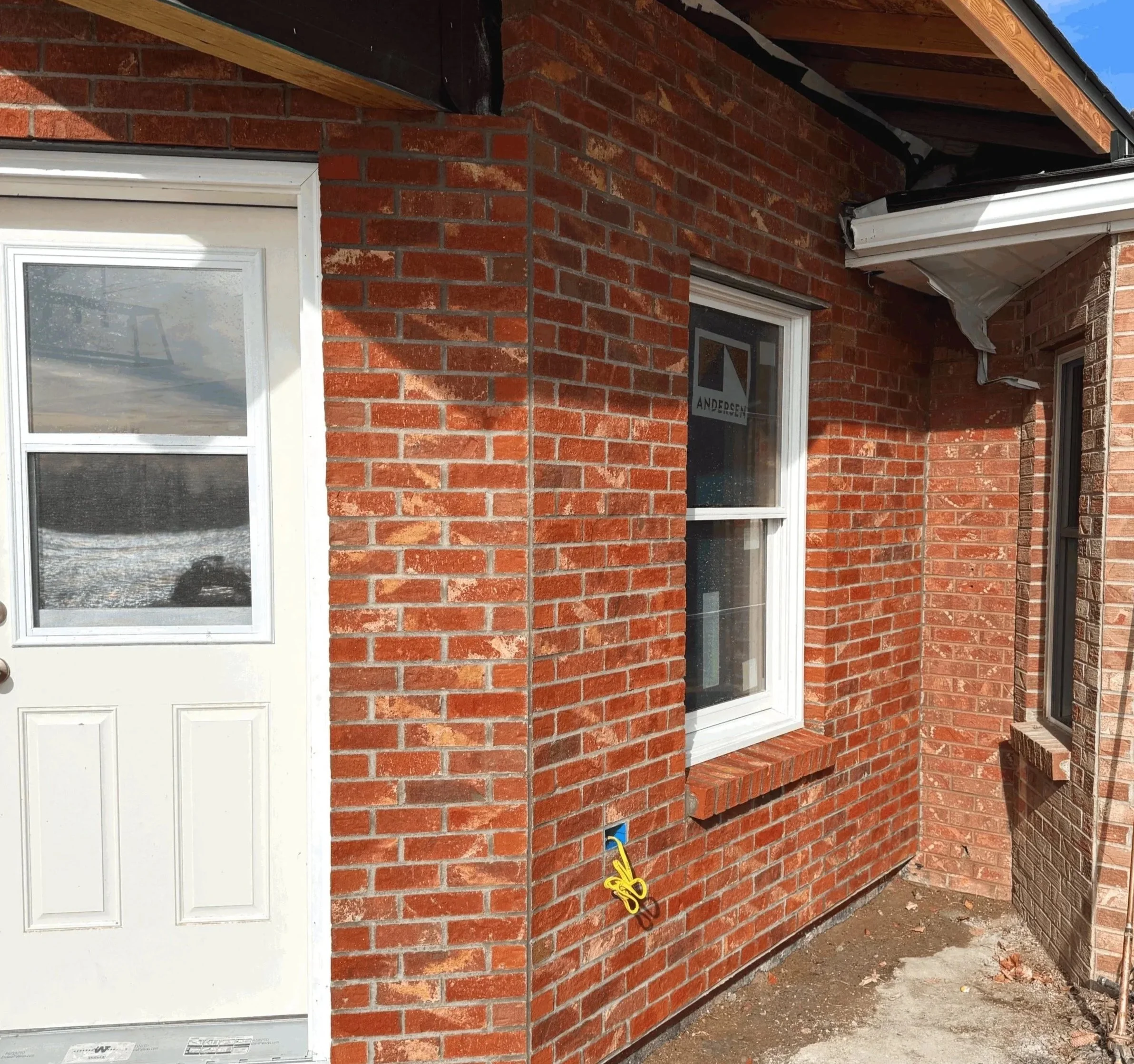 Close-up of a brick house exterior with a white door and window, showing electrical wiring and some construction materials near the foundation.