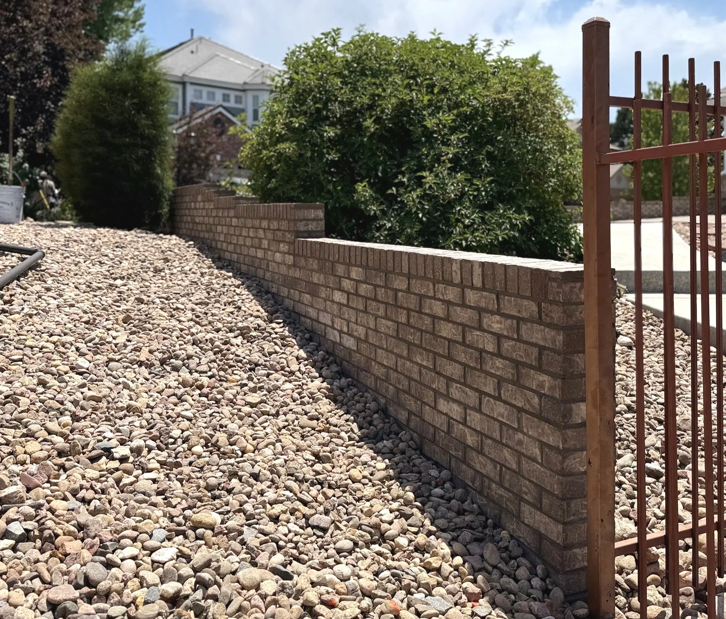 A residential yard with a brick wall, a metal gate, and decorative bushes surrounded by pebbles, with houses and trees in the background.