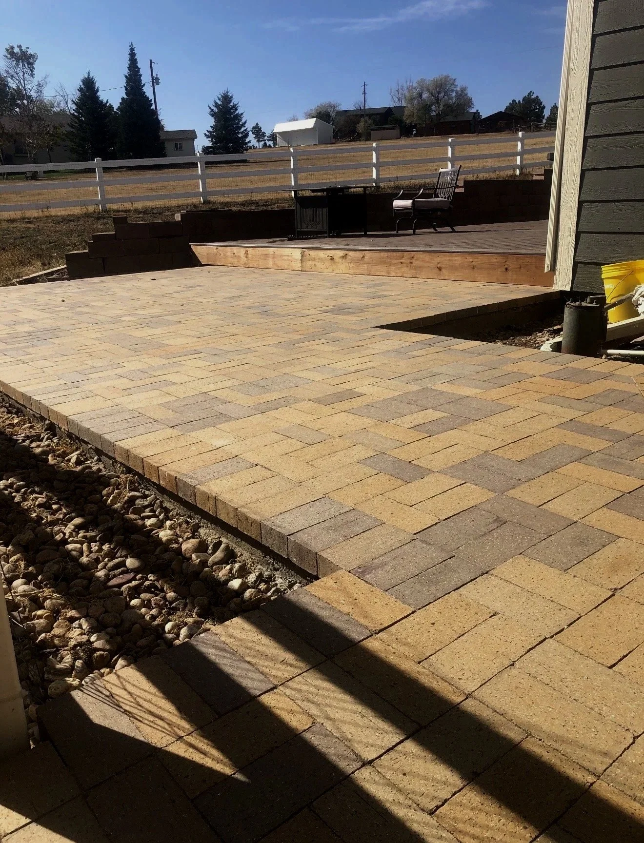 A newly paved stone patio with a step up to a raised wooden deck, outdoor furniture, and a white fence in a backyard during daytime.