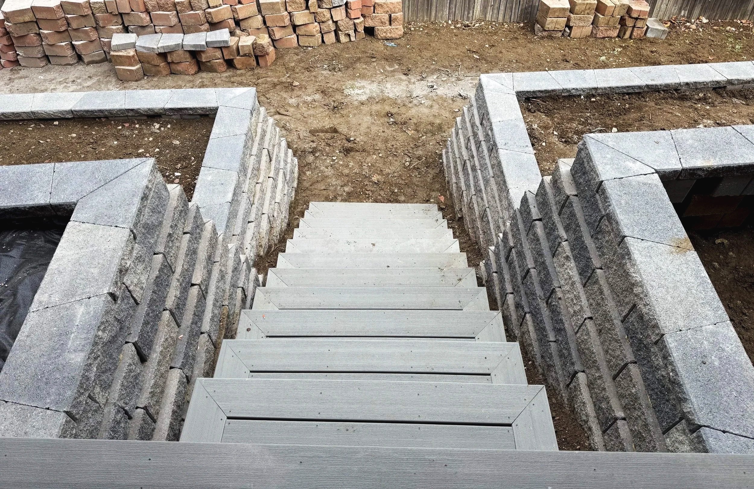 A set of outdoor stairs made of gray concrete leading down into a dirt area, with stone walls on either side and a dirt yard with bricks stacked against a wooden fence in the background.