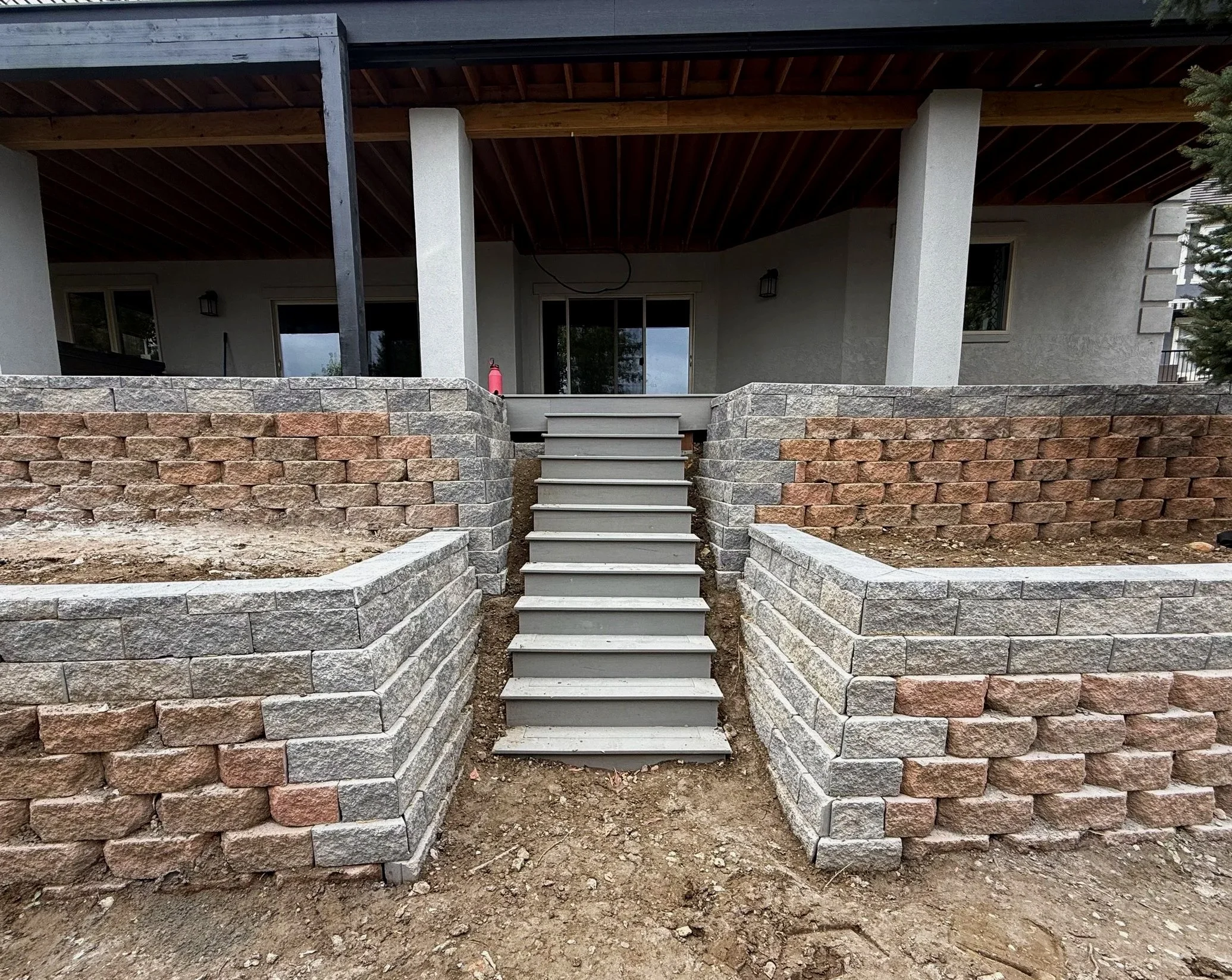 Image of an outdoor porch area with stairs leading up to a sliding glass door, supported by two concrete columns, and surrounded by a stone retaining wall. There is dirt ground in the foreground with some construction or landscaping work in progress.
