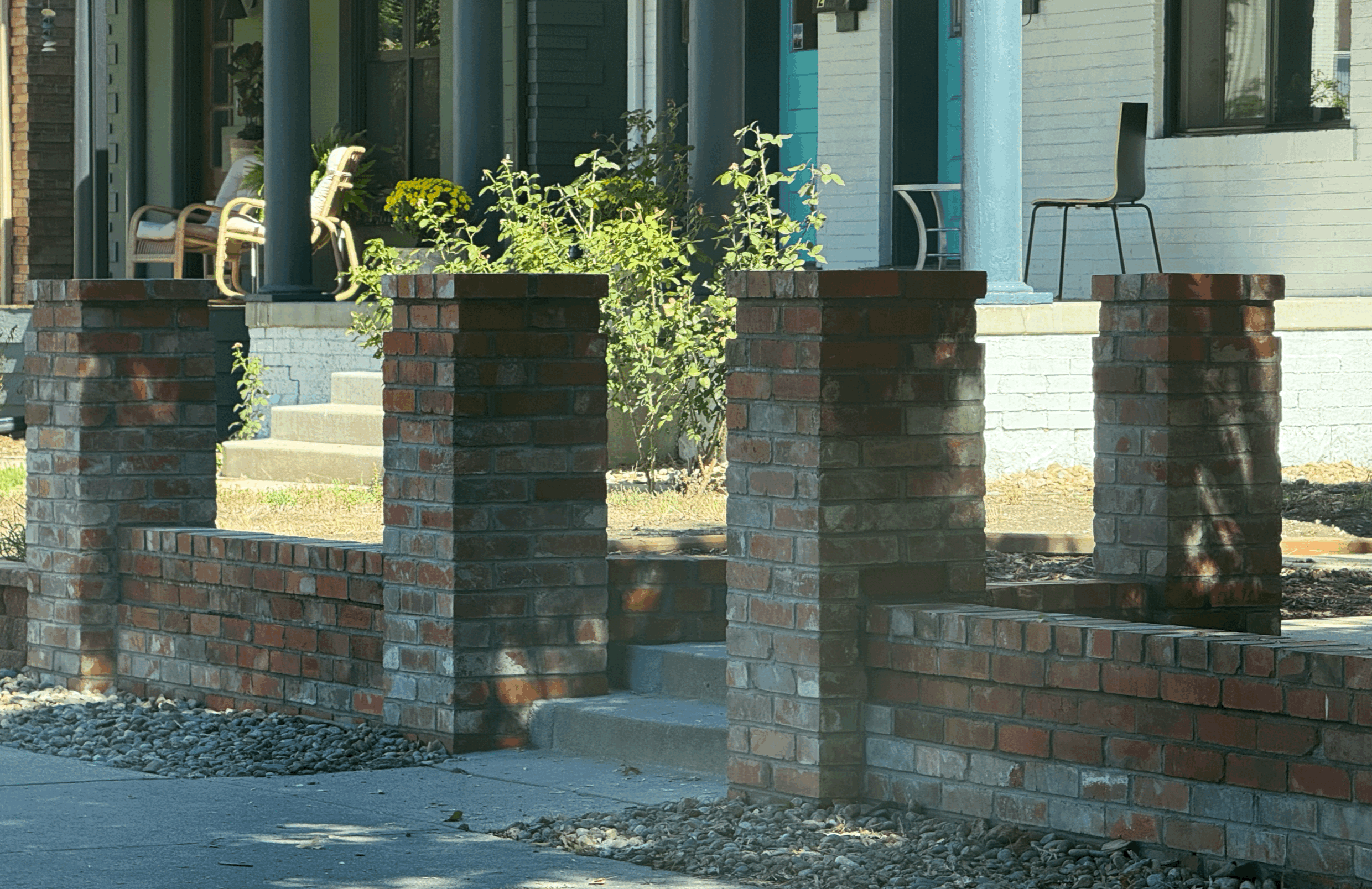 Front porch of a house with brick and white siding, decorated with potted plants, chairs, and a chair with a laptop, enclosed by a red brick low wall.