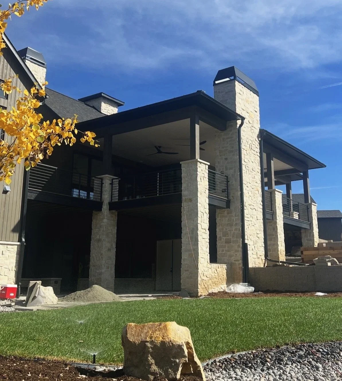 Under construction modern two-story house with stone and wood siding, covered porch, black railing, and a brick chimney on a sunny day, with a green lawn and a rock in the foreground.