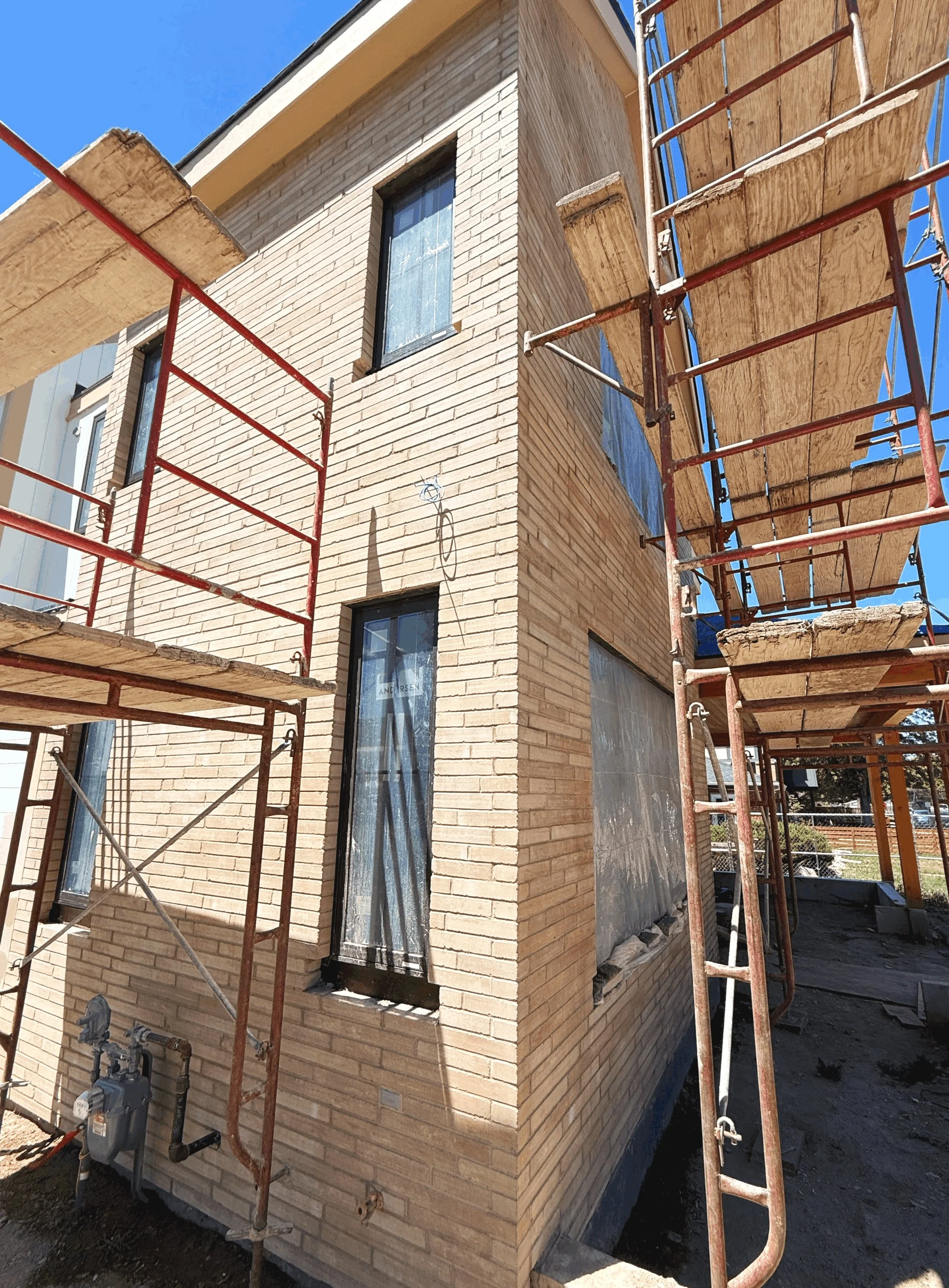 Construction site with scaffolding around a brick building, with visible windows, pipes, and a clear blue sky.