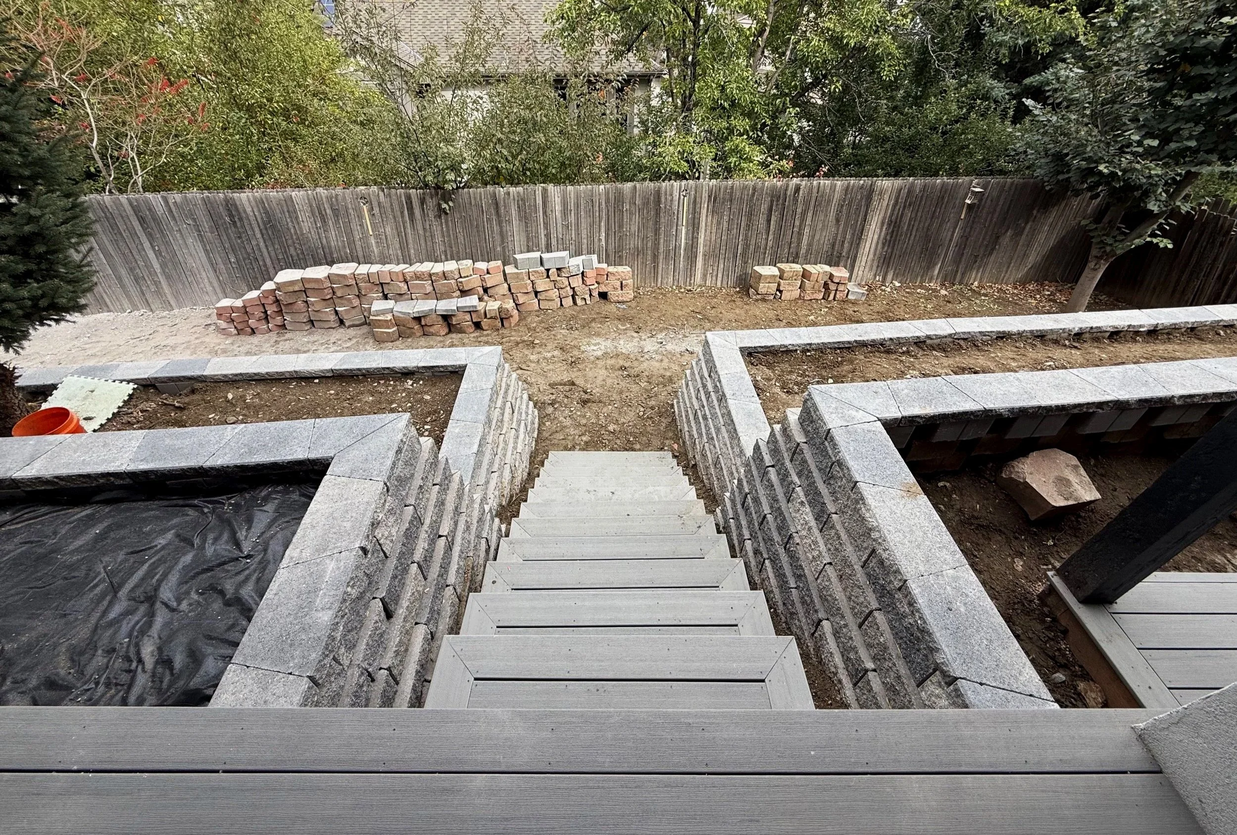Wooden stairs leading down to a yard with a brick border. A wooden fence and trees are in the background.