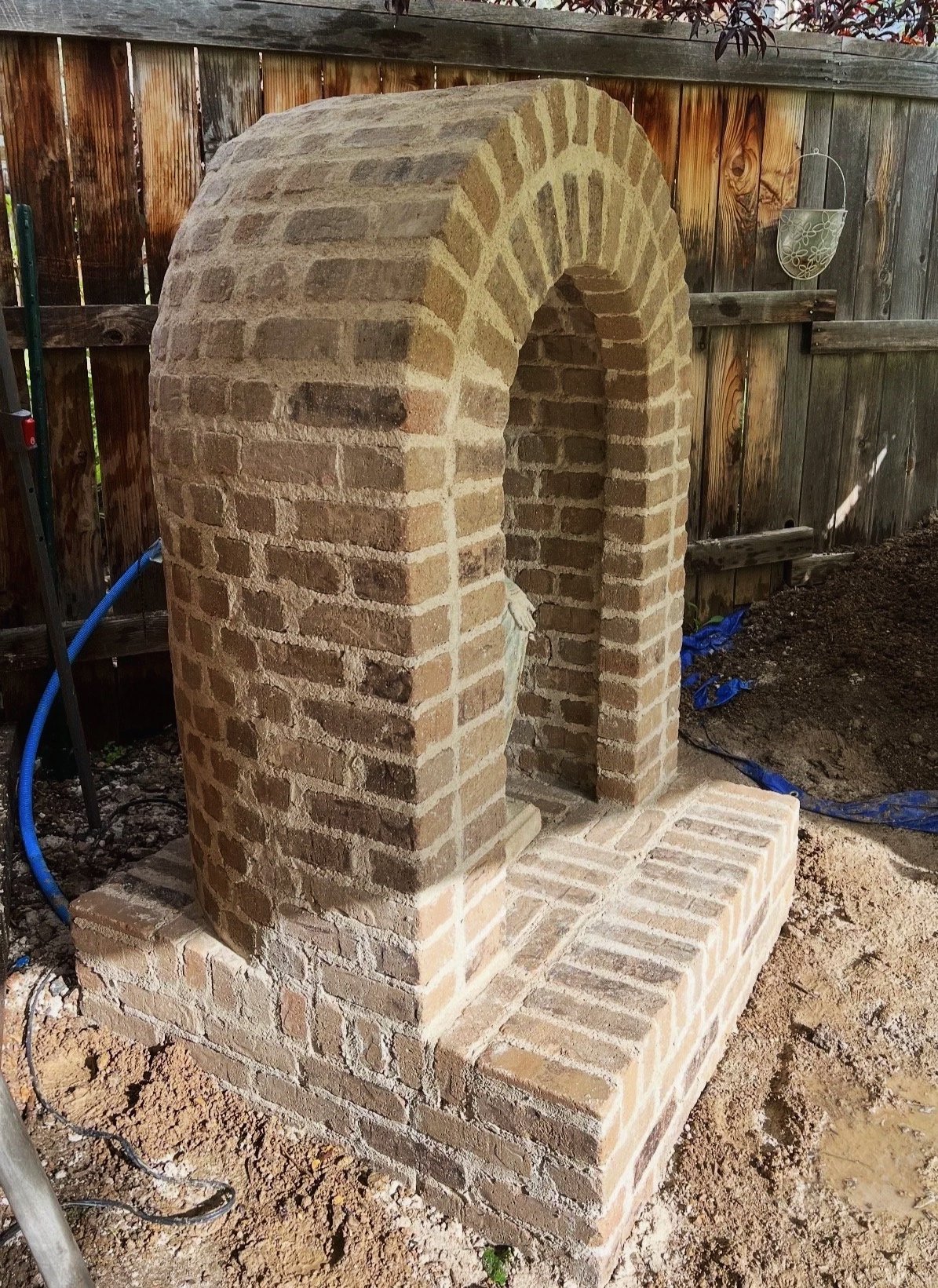 A brick outdoor fireplace or oven under construction in a backyard, with a curved brick arch and brick base, surrounded by dirt and construction materials.